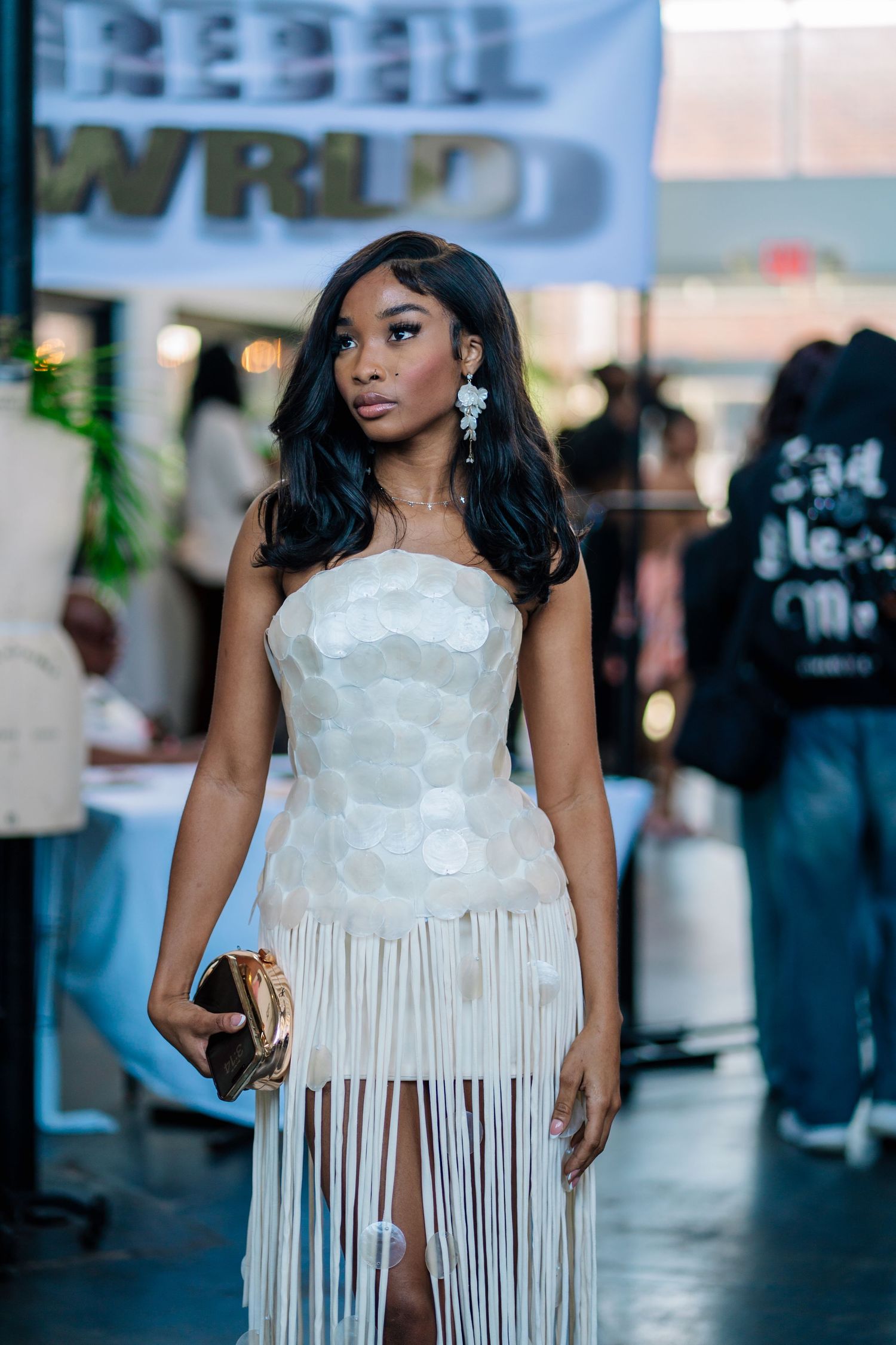 Young woman in white fringed strapless dress with floral bodice, holding gold clutch at fashion event.