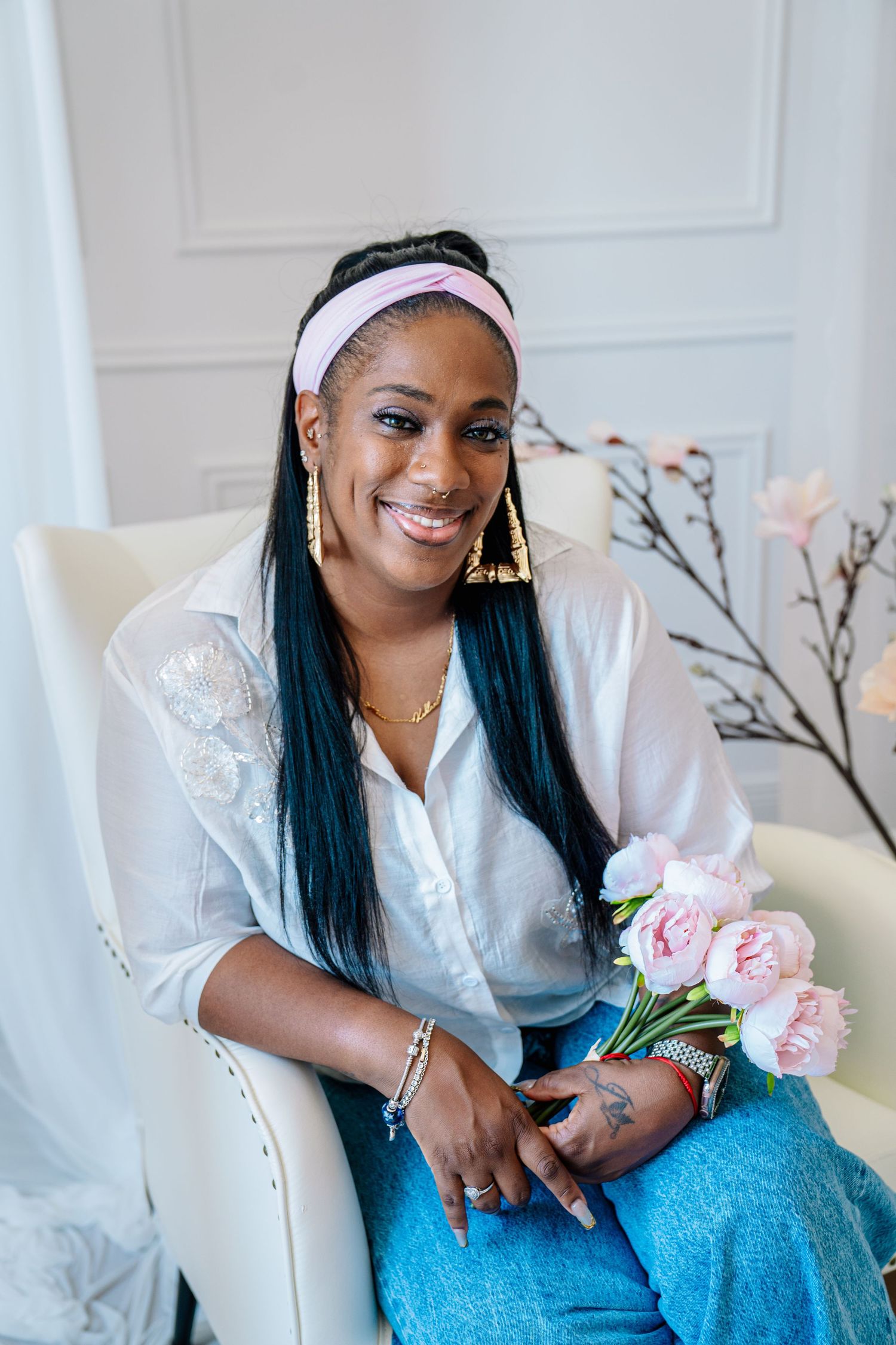 Smiling Black woman with blue braids and pink headband sits in white chair holding pink peonies wearing jeans and white shirt.