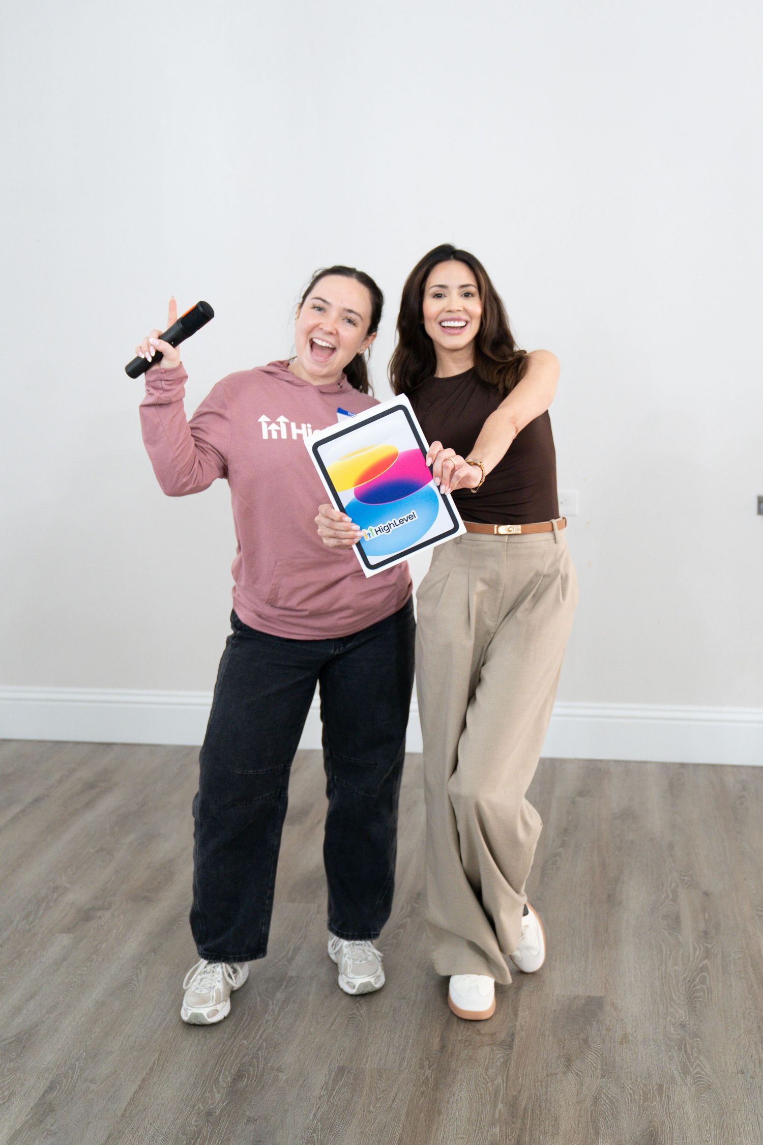 Two women smiling and posing with an iPad and microphone against a white wall on hardwood floors.