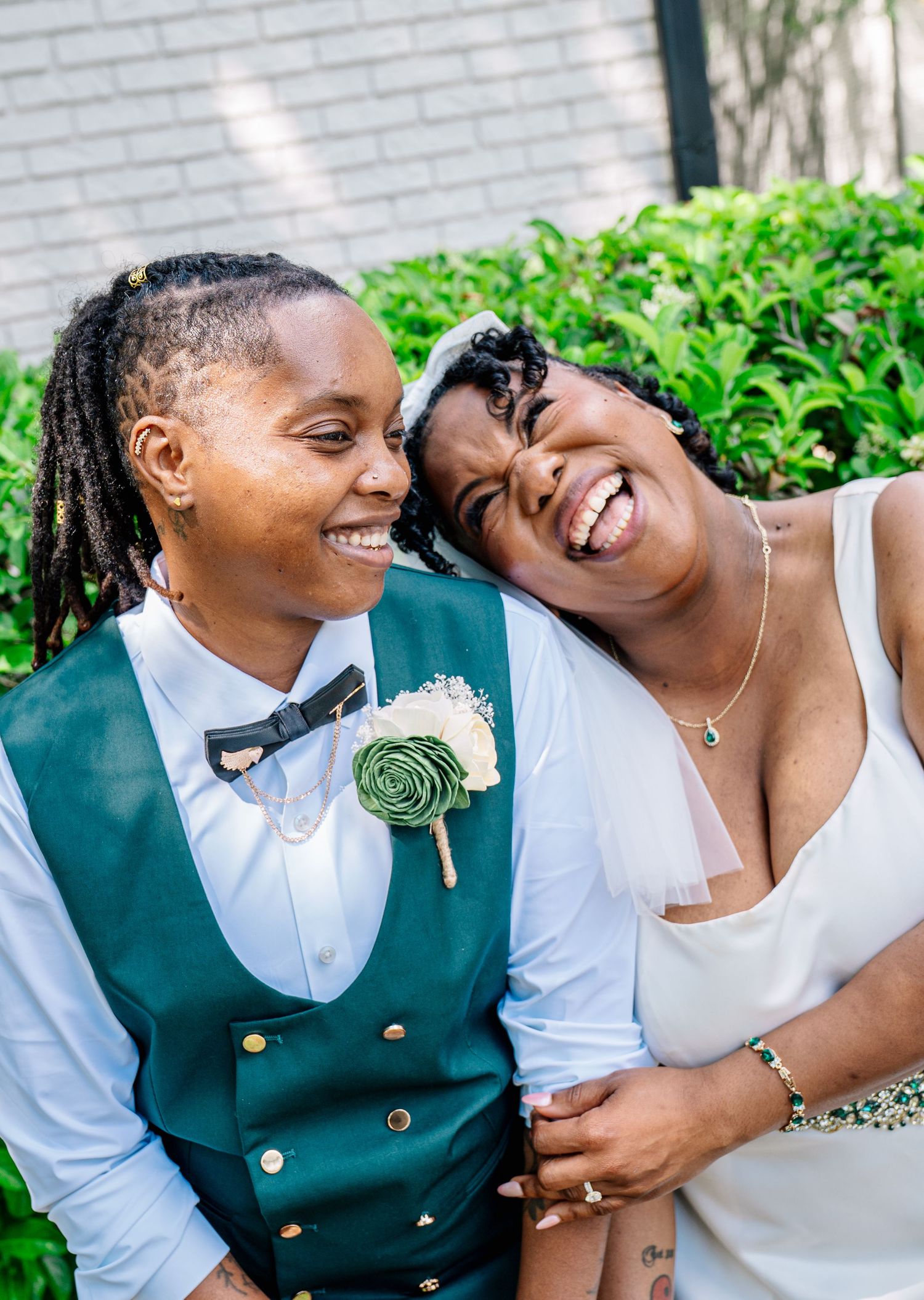 Two brides laughing joyfully outdoors, one in a green vest with boutonniere, the other in a white wedding dress.