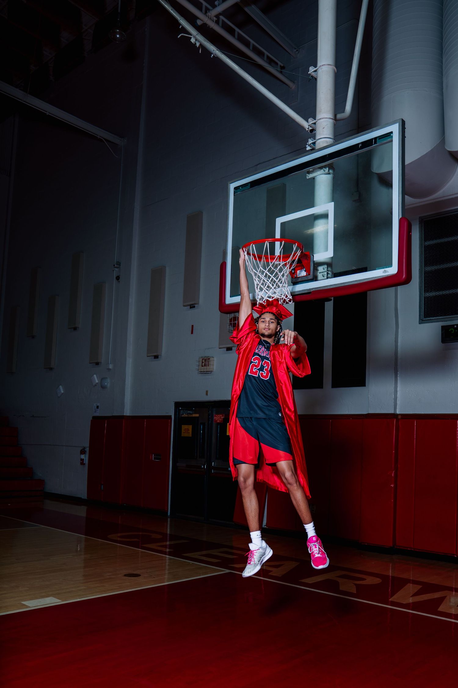 Basketball player in red jersey number 23 leaping toward hoop in indoor gym, wearing red shorts and pink sneakers.