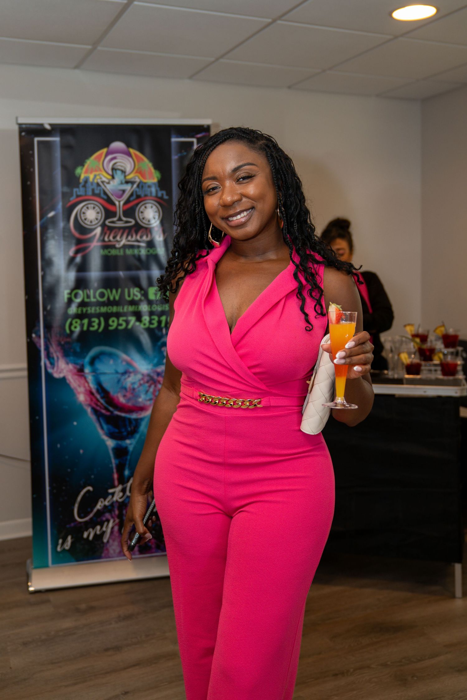 Woman in pink jumpsuit smiling and holding a cocktail at a social event with a bar banner in the background.