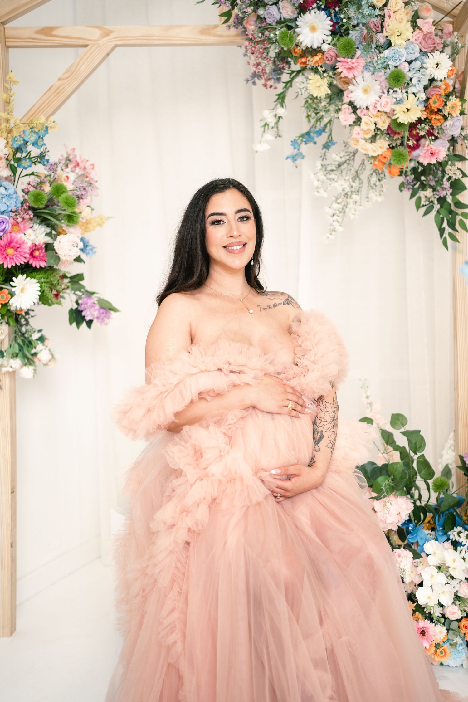 Pregnant woman in elegant pink tulle off-shoulder gown poses under colorful floral arch in studio setting.