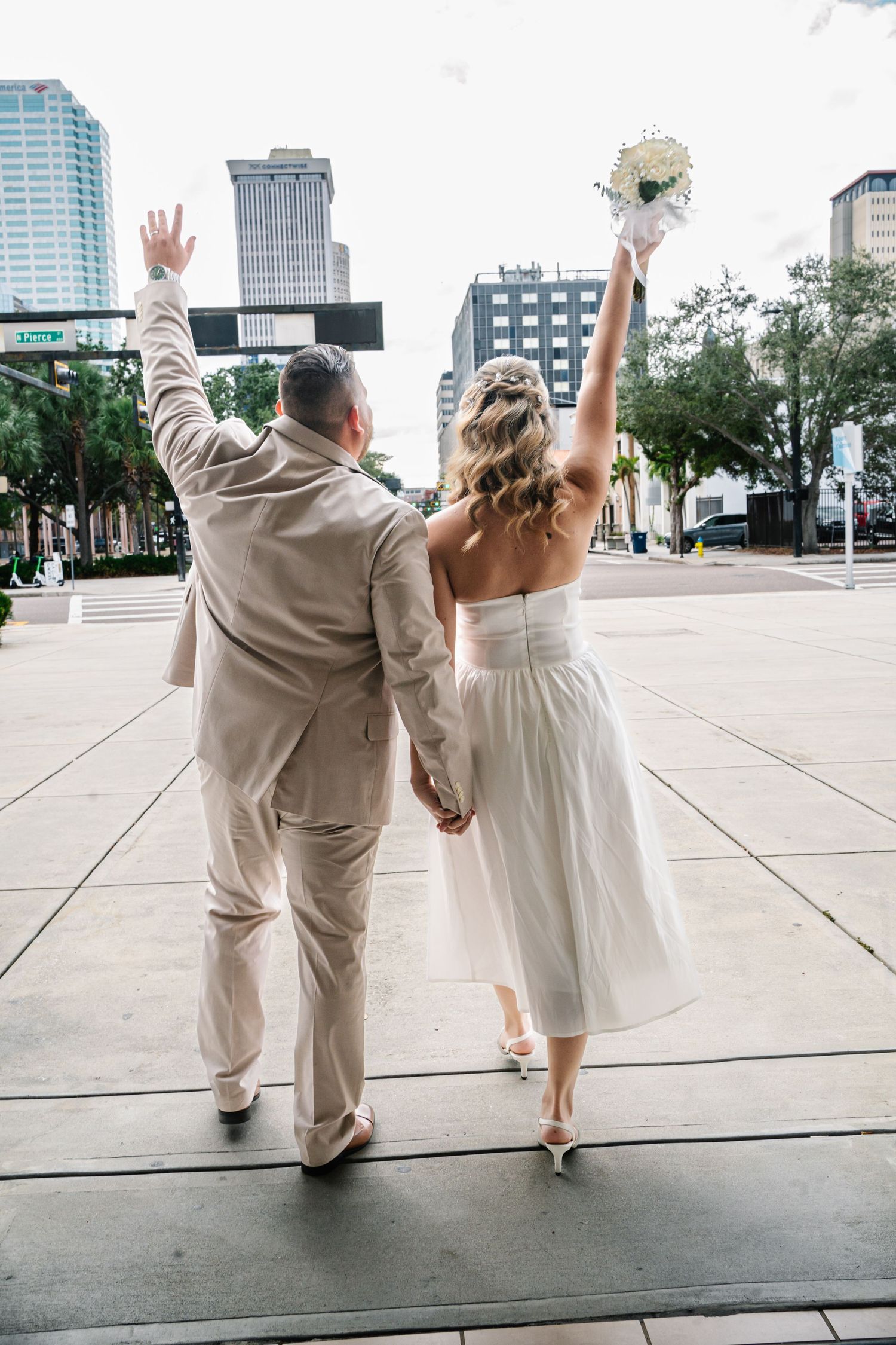 A couple in light-colored wedding attire poses playfully on an urban plaza with city buildings in the background.
