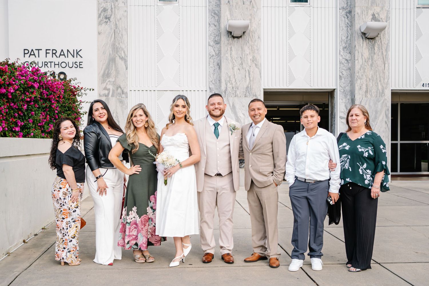 Wedding party poses outside Pat Frank Courthouse, bride in white dress, groom in tan suit, surrounded by smiling family members.