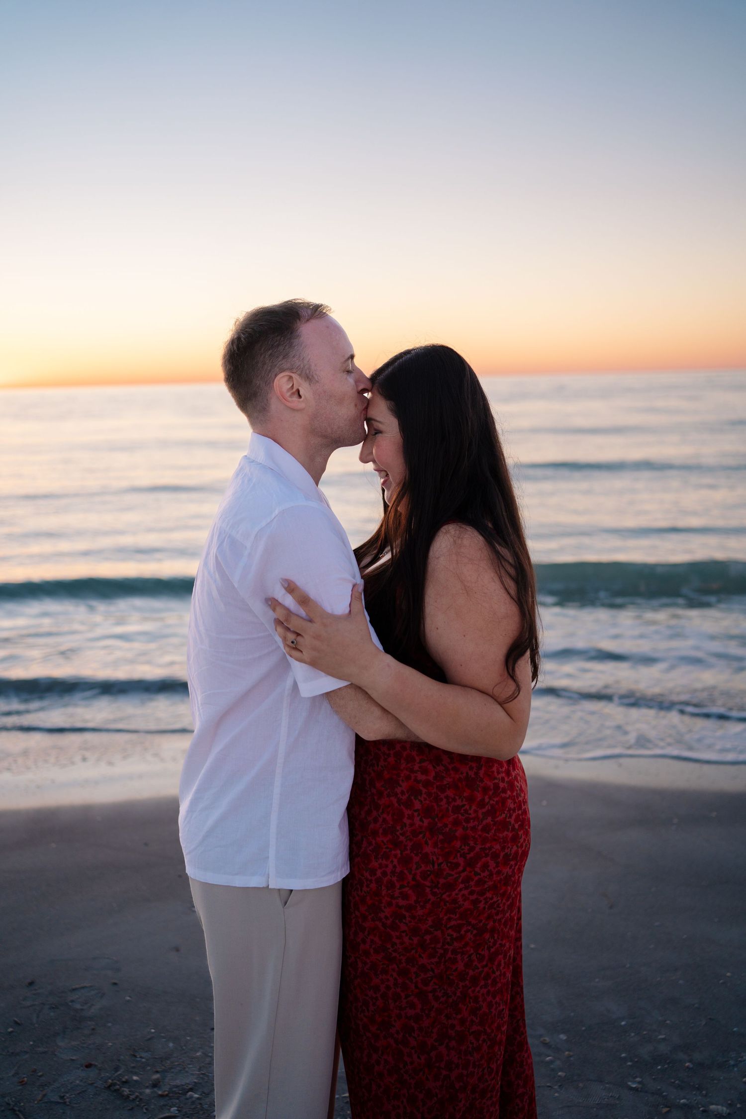 Couple embracing and kissing at sunset on a beach, woman in red dress, man in white shirt, ocean backdrop.