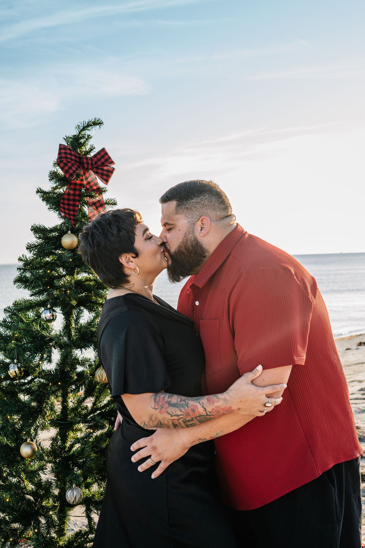 Couple kissing beside a decorated Christmas tree on a beach, man in red shirt and woman in black dress.