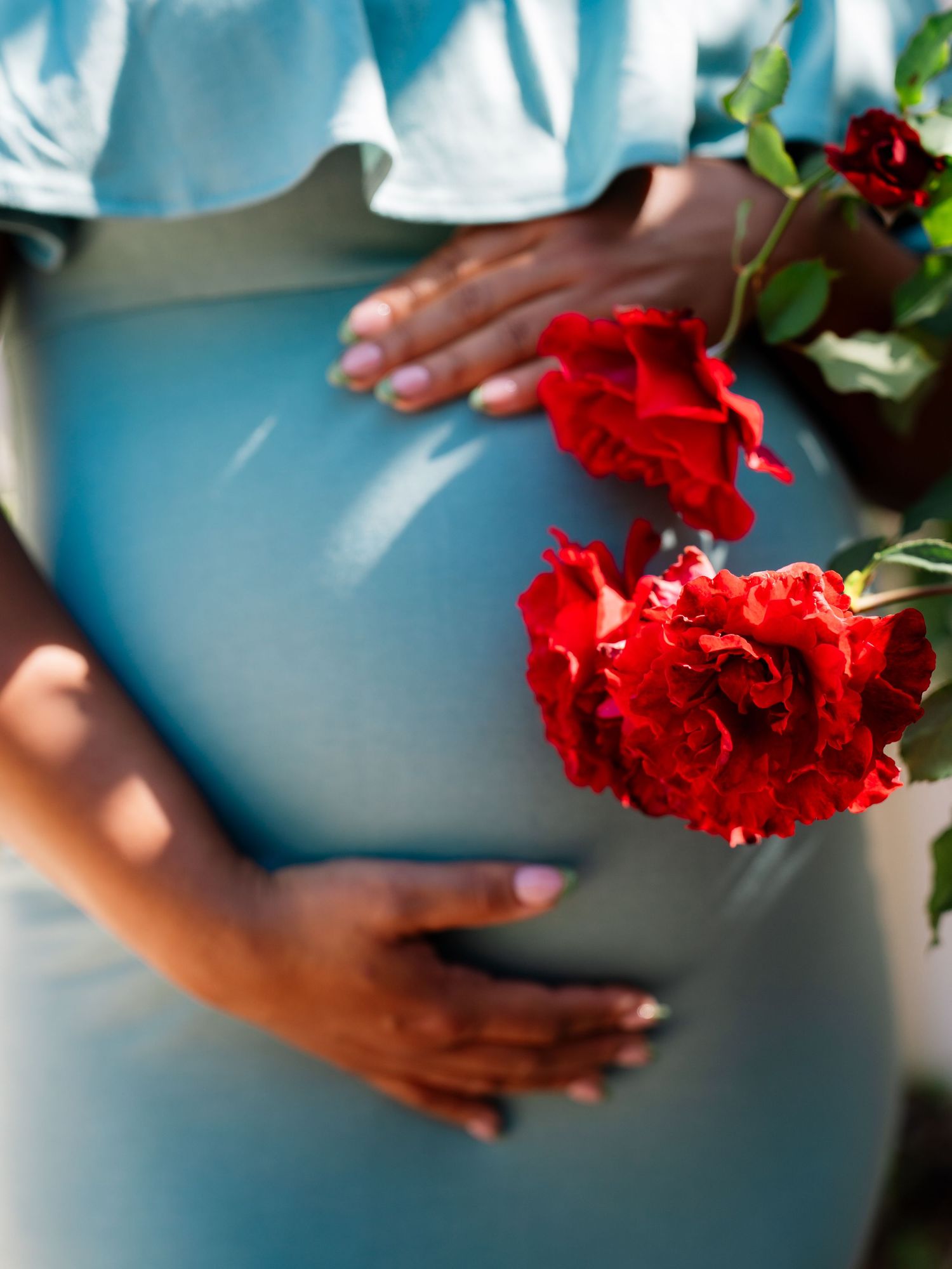 Pregnant woman in light blue dress holding red roses, cradling baby bump with manicured hands outdoors.