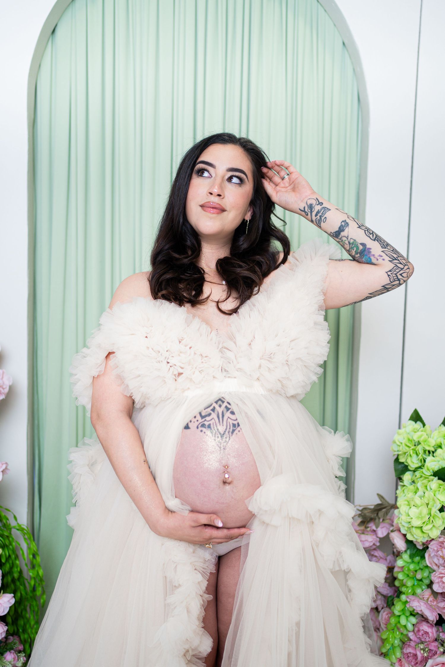 Pregnant woman in white ruffled maternity gown with tattoos, cradling baby bump, posing in elegant floral studio setting.