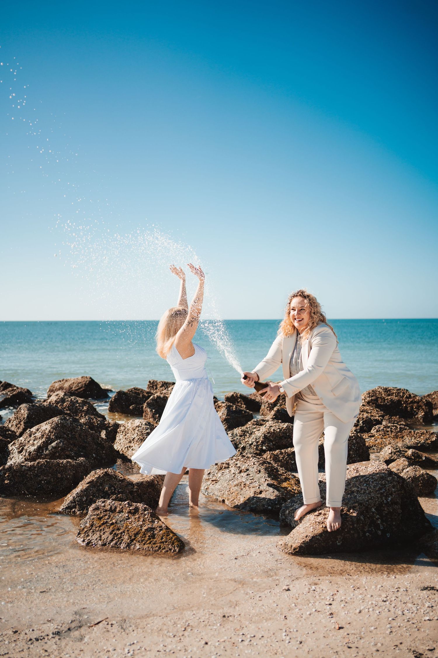 Couple celebrating on rocky beach, woman in white dress tossing champagne, man in beige suit, blue ocean backdrop.