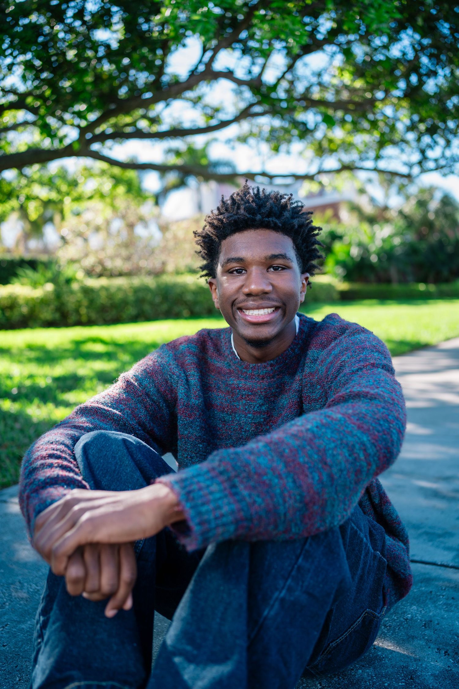 Young Black man smiling outdoors, sitting on pavement in a colorful sweater and jeans, with green trees in background.