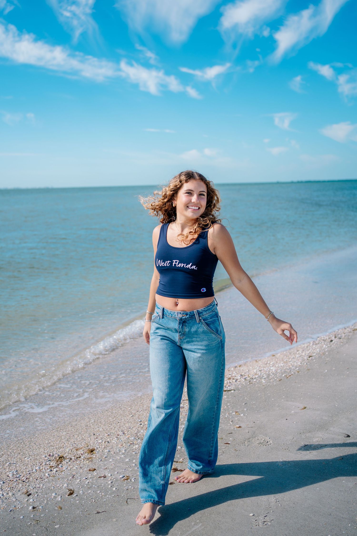 Young woman in a navy crop top and jeans smiling on a sunny Florida beach with blue ocean waves.