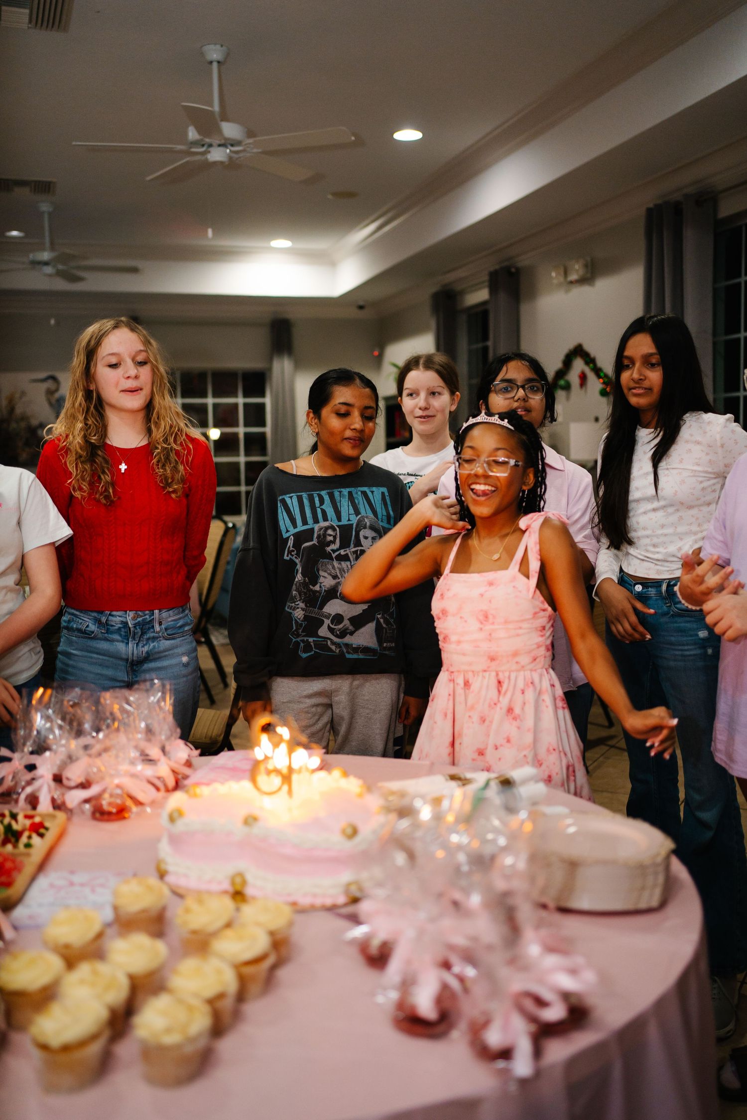 A joyful birthday girl in a pink dress smiles before her cake surrounded by friends at an indoor celebration.