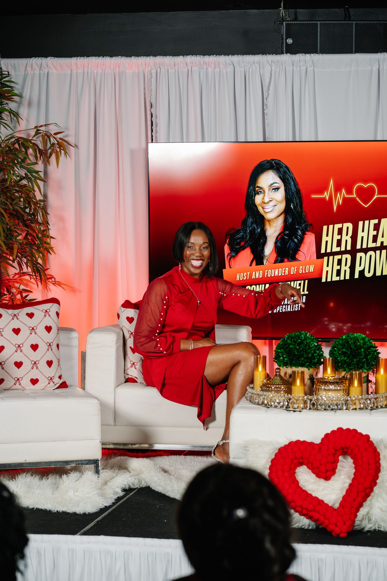 Woman in red dress poses on white sofa at Her Heart Her Power event with decorative heart display backdrop.