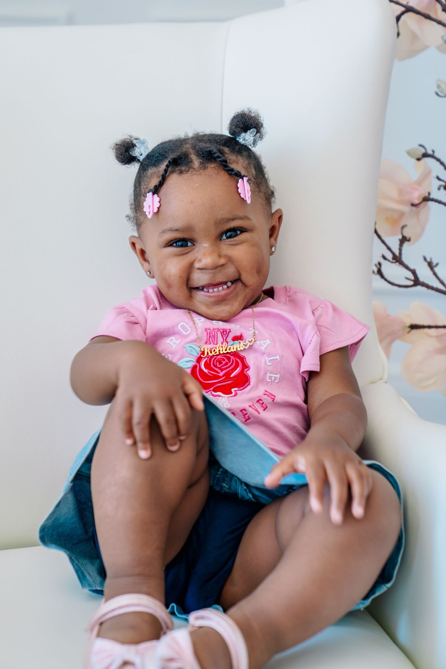 Adorable smiling toddler girl with pigtails wearing a pink shirt and denim skirt sitting on a white chair.