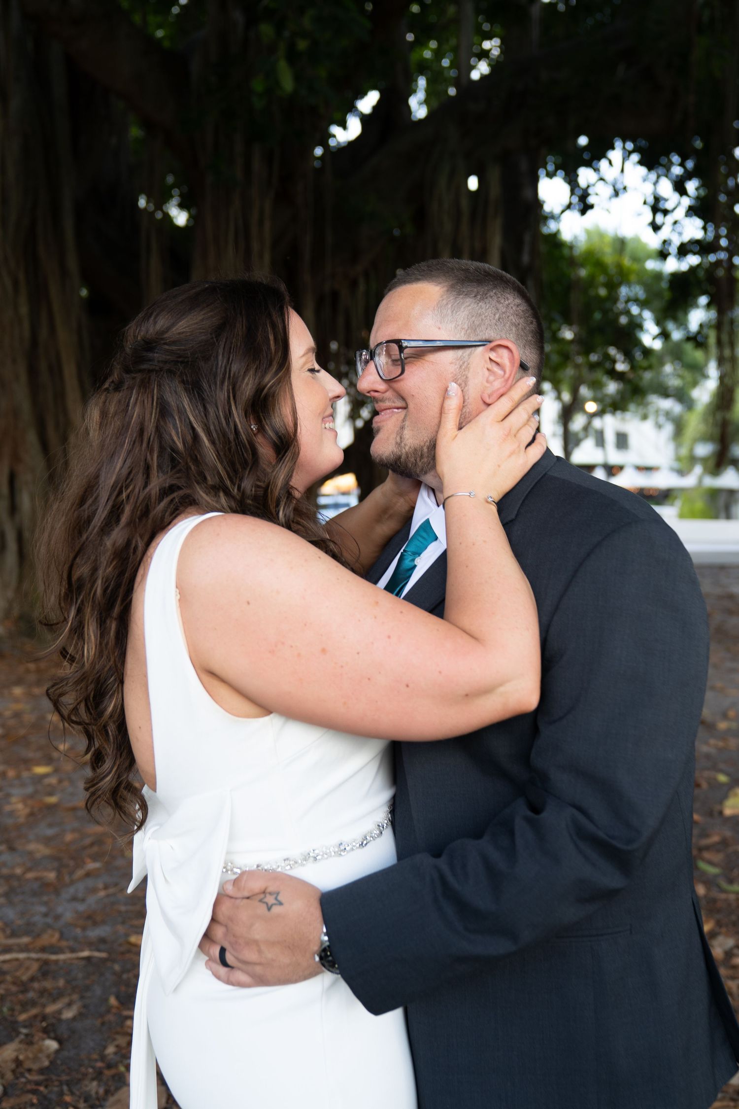 Bride and groom share a romantic embrace, smiling at each other near a large banyan tree outdoors.
