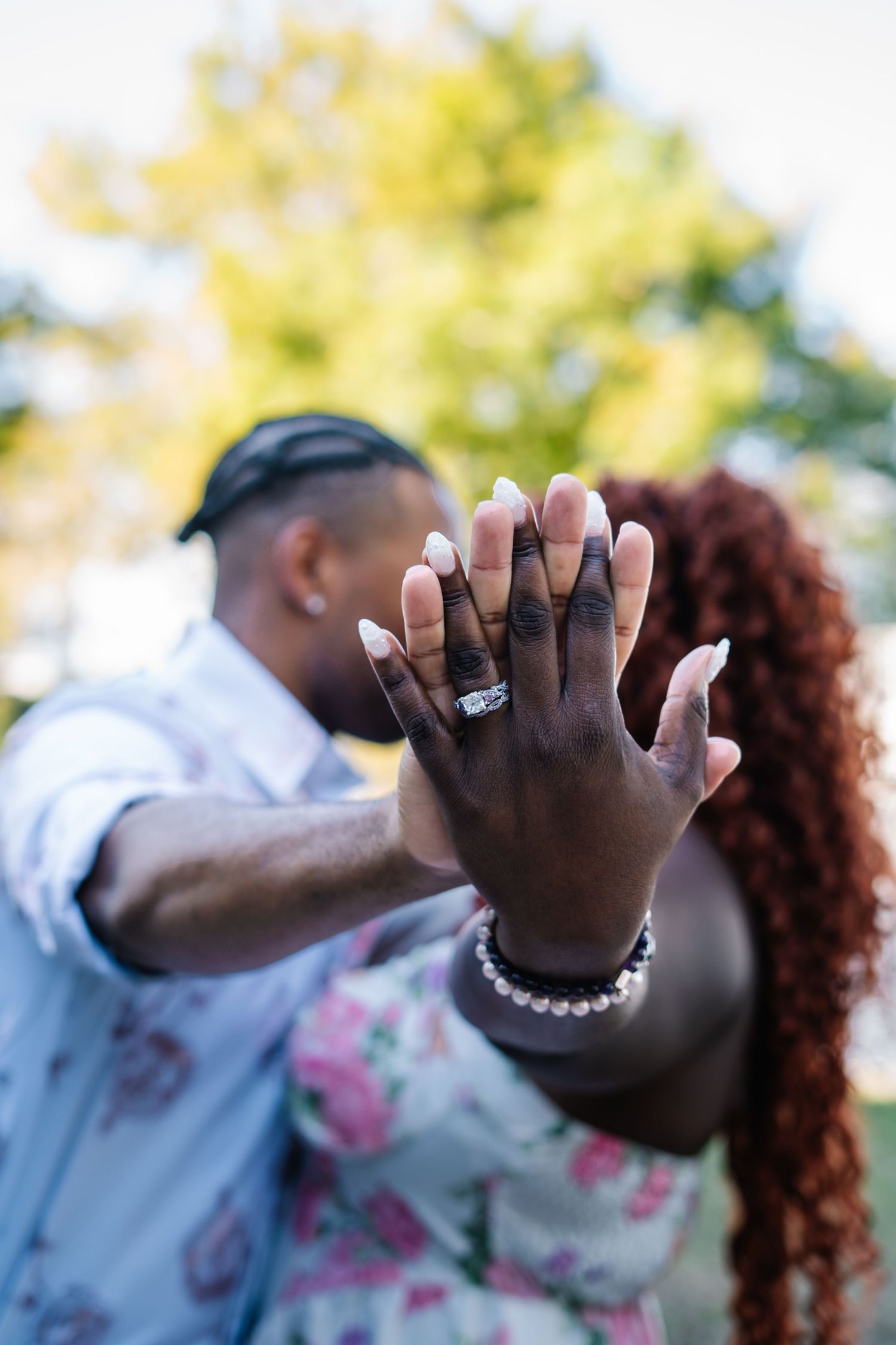 Couple showing off engagement ring outdoors, hands raised toward camera with bokeh green trees background.