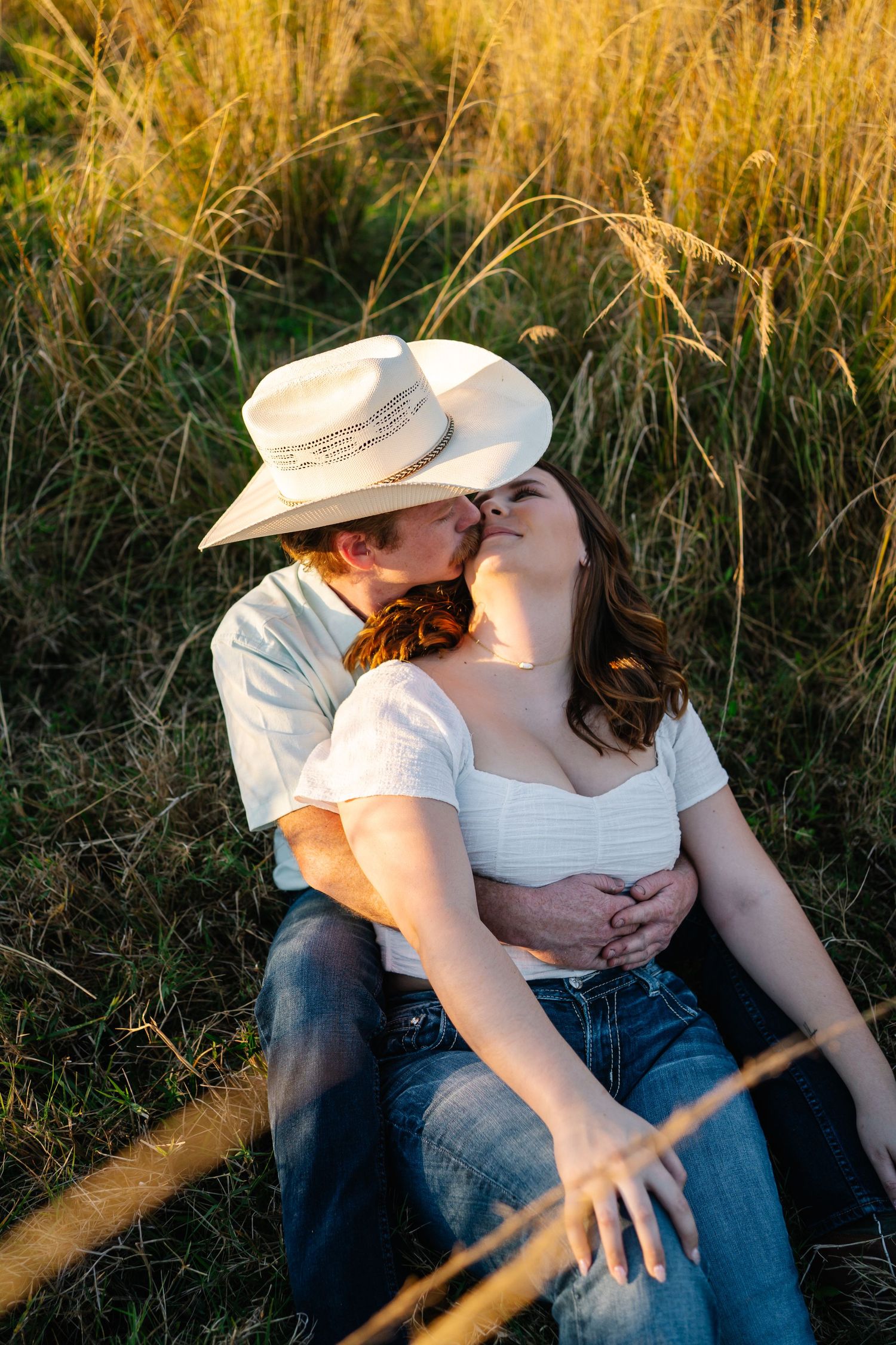 Couple sharing a romantic kiss in a golden field at sunset, man wearing a white cowboy hat and woman in a white crop top.