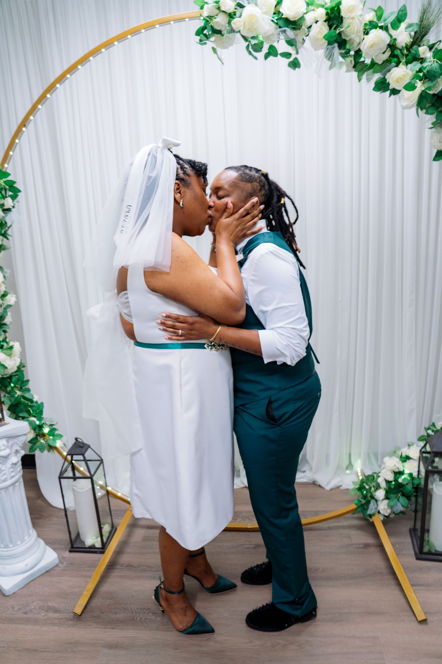 Two brides sharing a first kiss at the altar under a floral arch during their wedding ceremony.