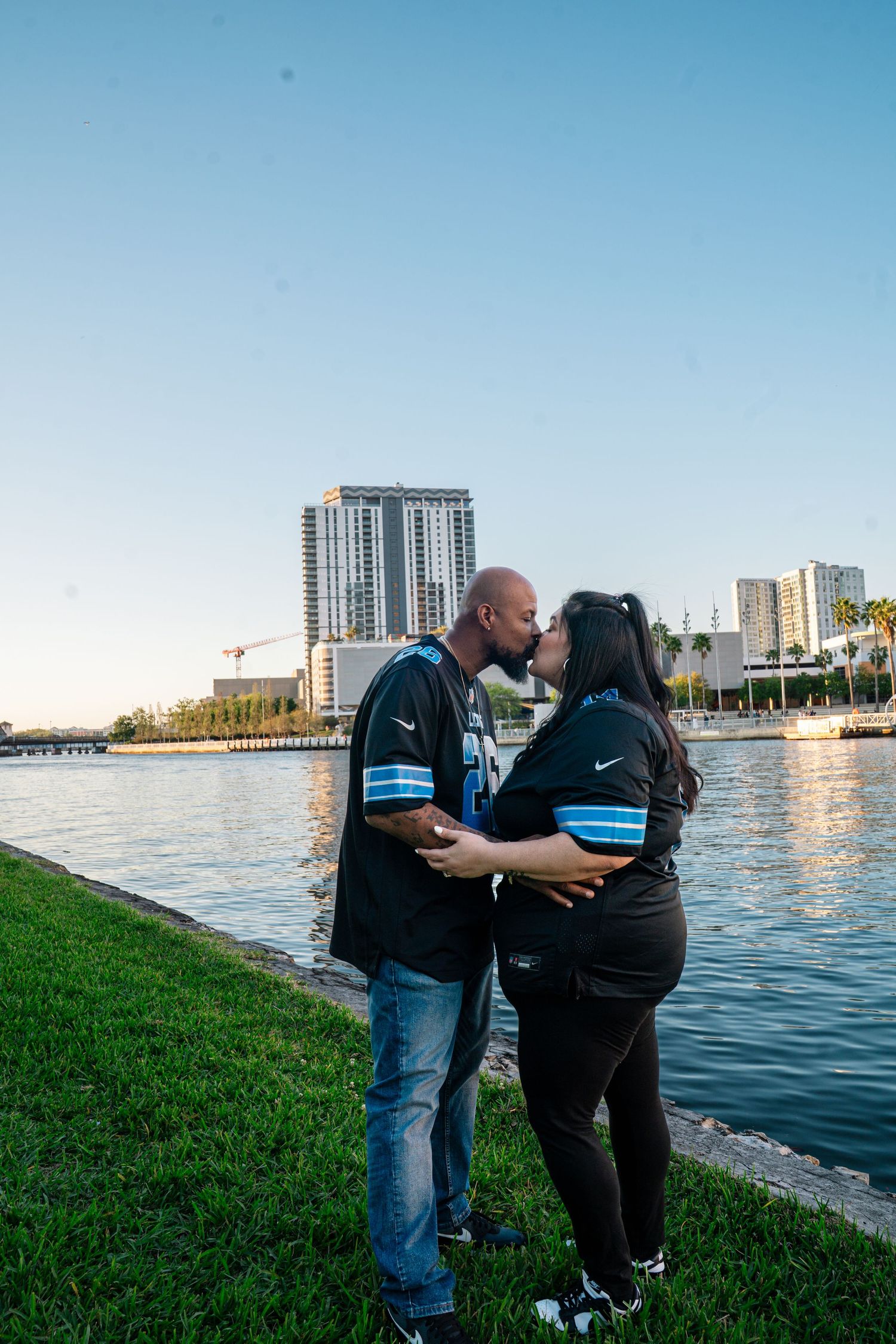 Couple kissing by a waterfront park wearing matching black Nike jerseys with a city skyline at sunset.