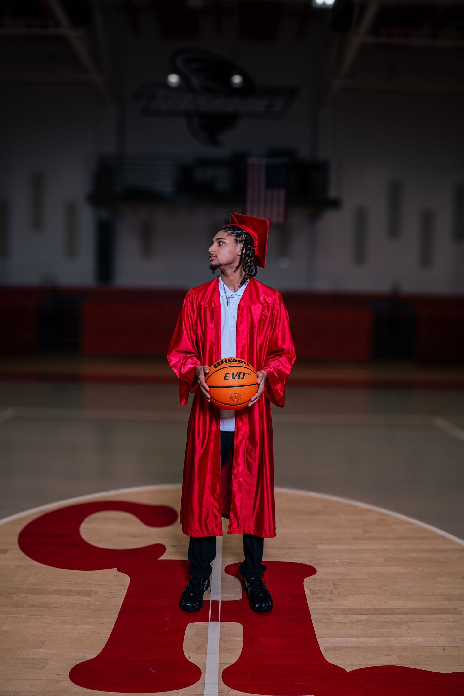 A young man in a red graduation gown and cap holds a basketball on a gym court center logo.