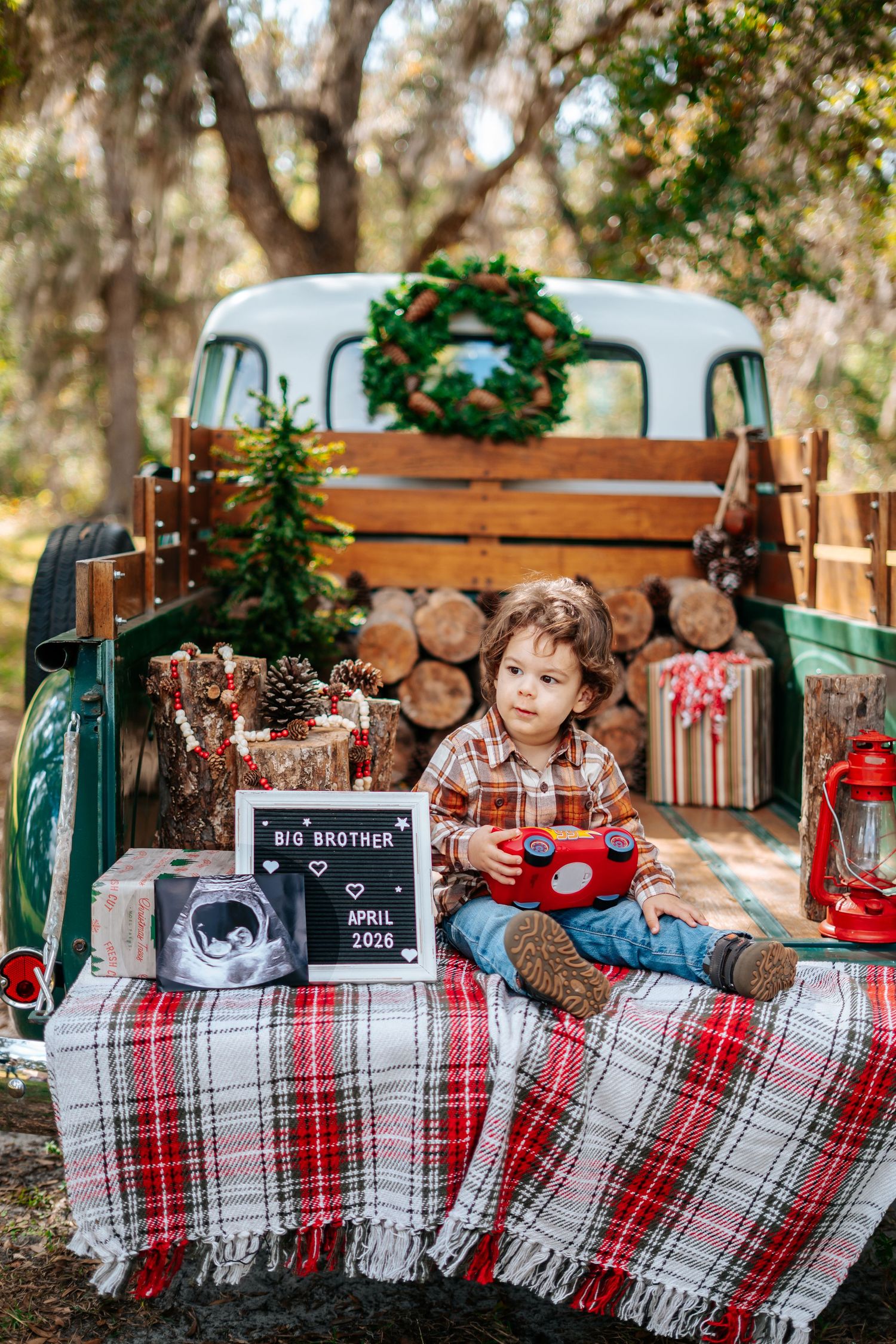 A young boy sits in a vintage truck bed with Christmas decor and a Big Brother pregnancy announcement sign for April 2026.
