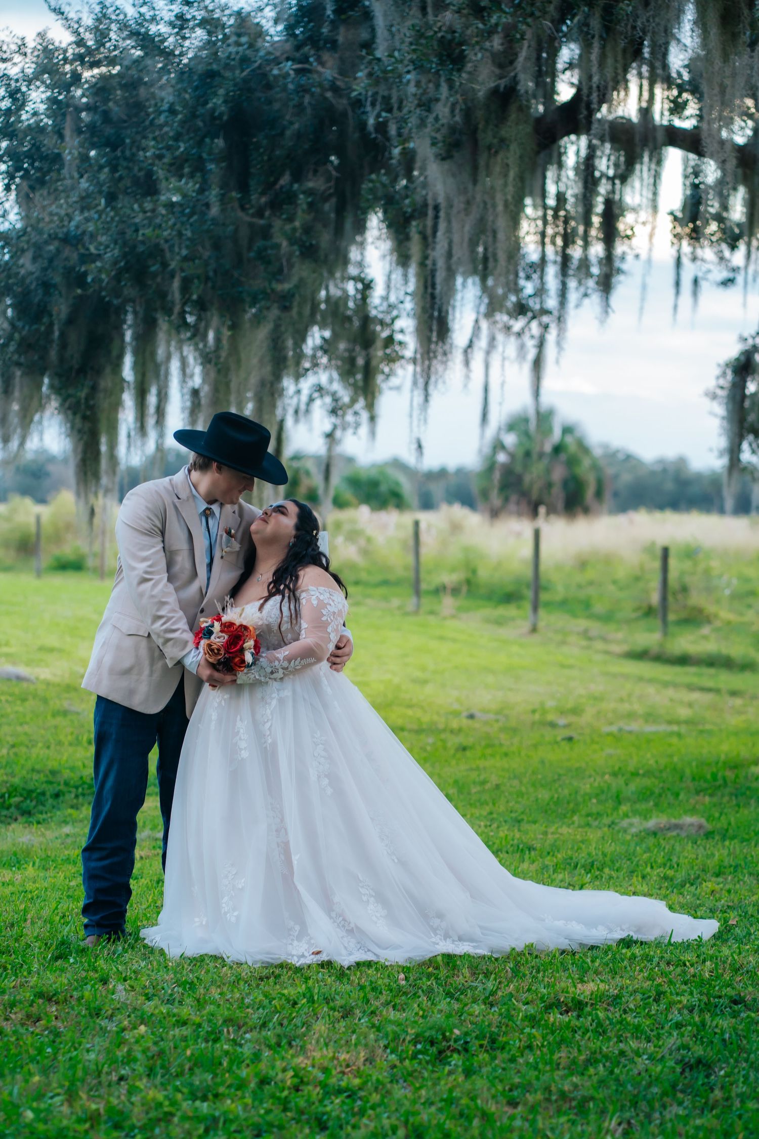 Wedding couple embracing on grassy field with Spanish moss draped trees creating romantic backdrop.