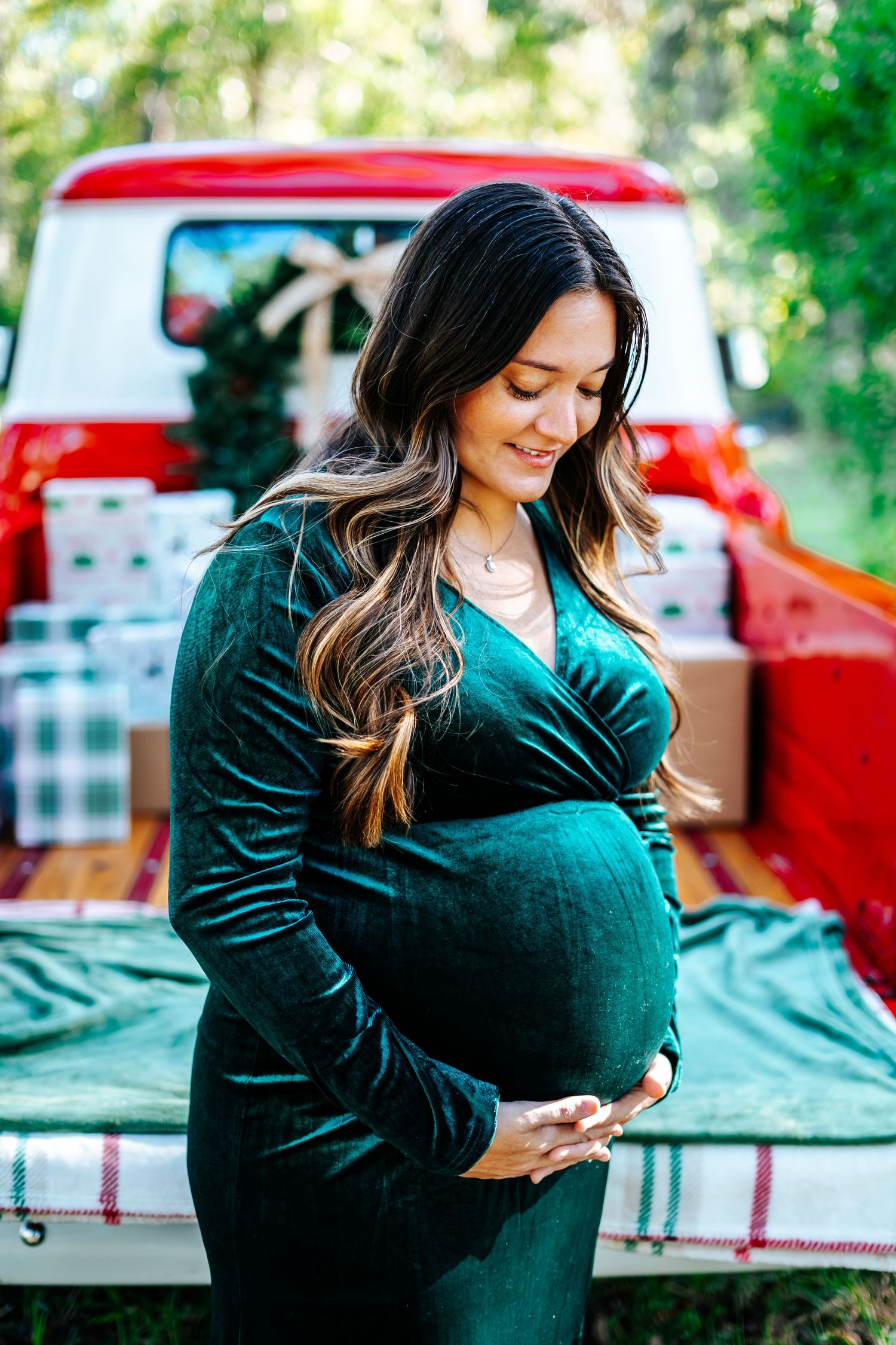 Expectant mother in emerald velvet dress poses by vintage red pickup truck with blanket in truck bed.