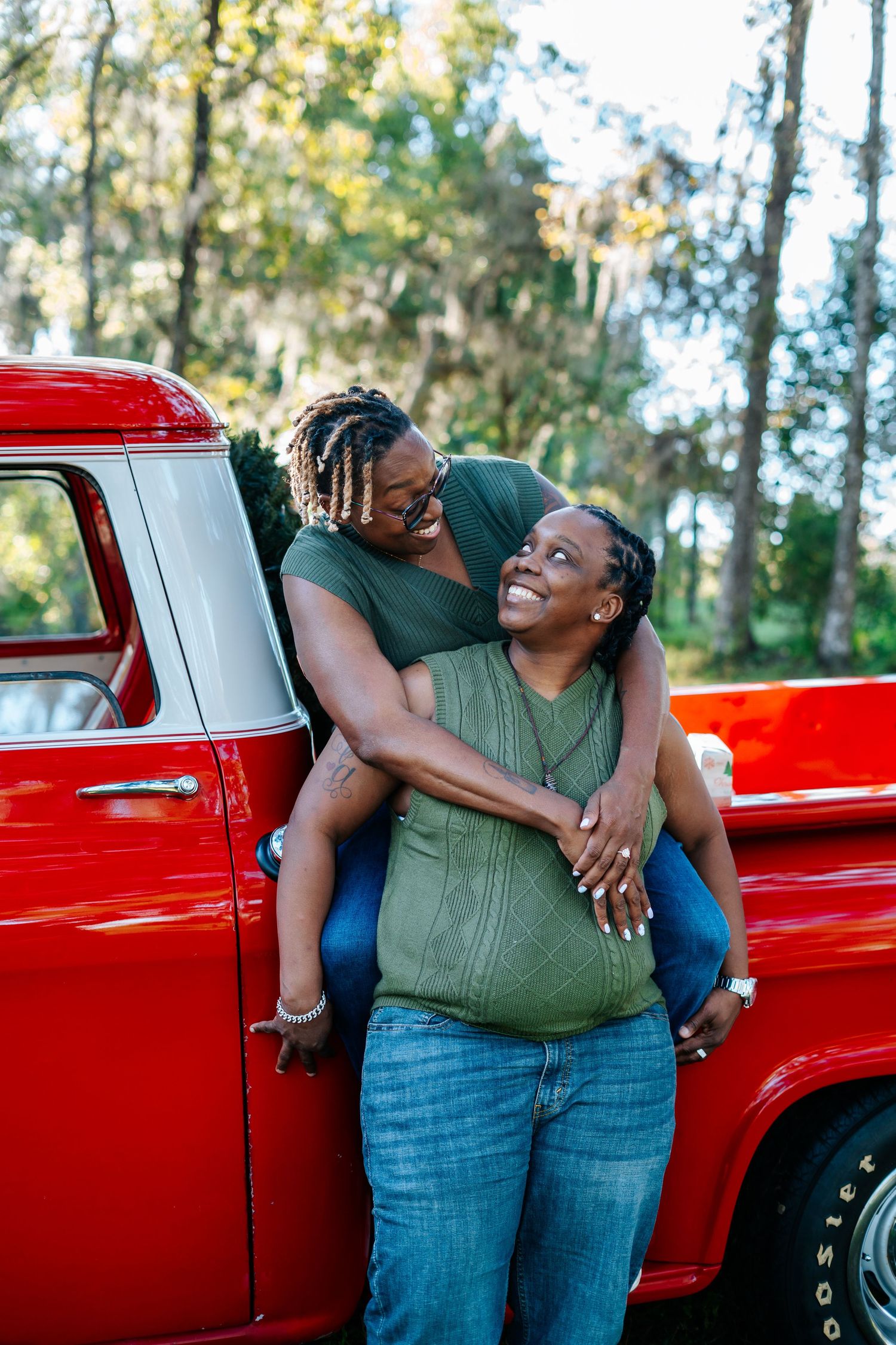 A couple shares a joyful embrace while leaning against a vintage red pickup truck in a wooded outdoor setting.