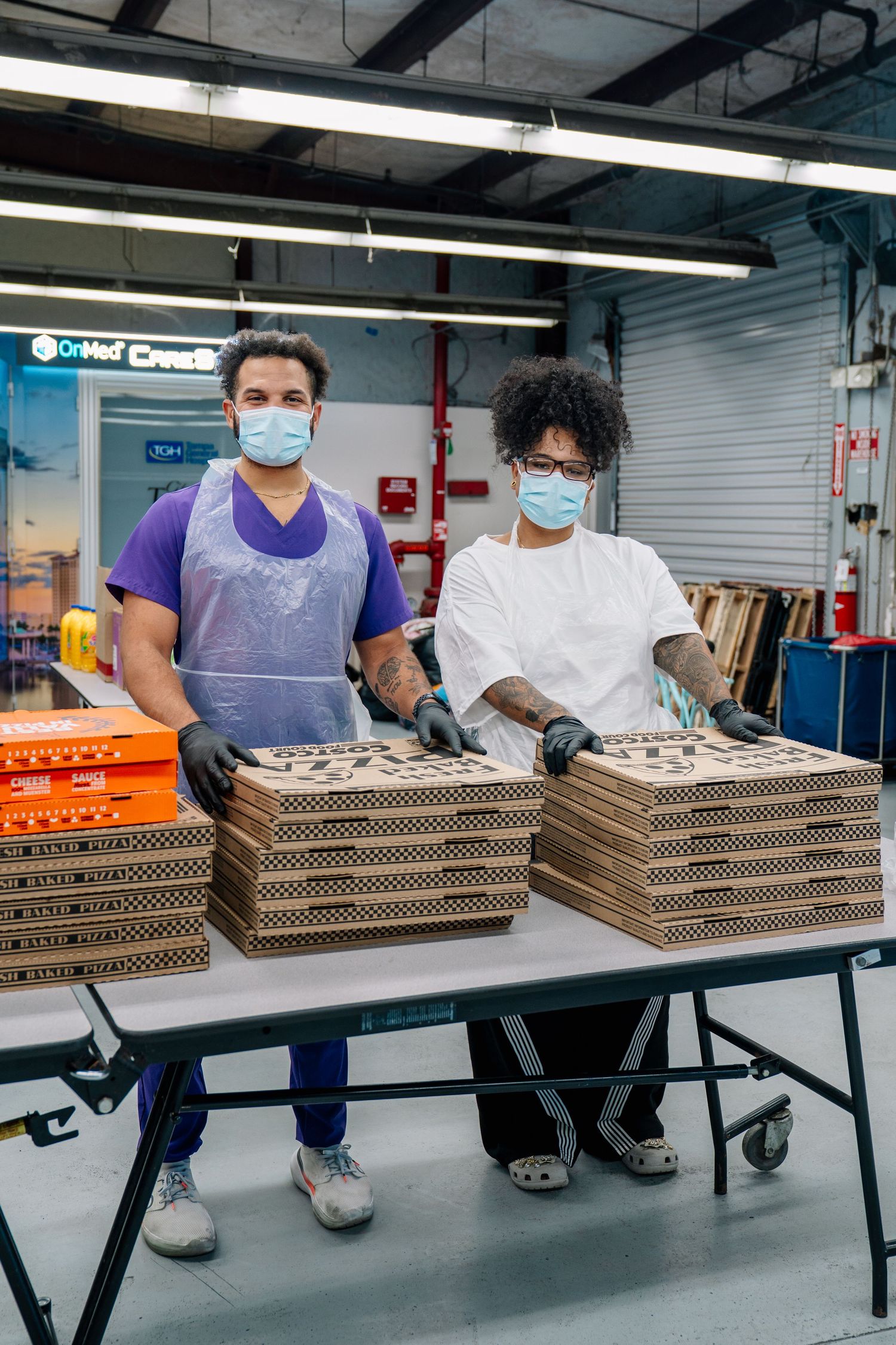Two workers in masks and gloves stack cardboard pizza boxes on a table in a warehouse setting.