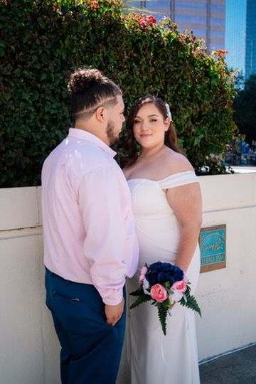 A couple shares an intimate moment against a white wall with flowering bushes and blue buildings in the background.
