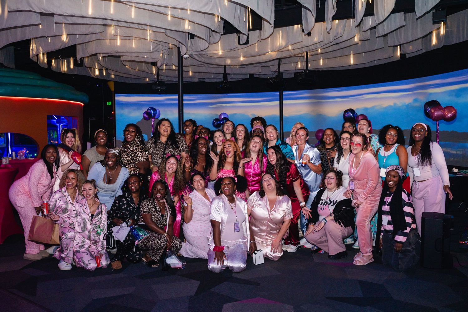 Large group of women in pajamas and robes posing together at a colorful indoor event venue.
