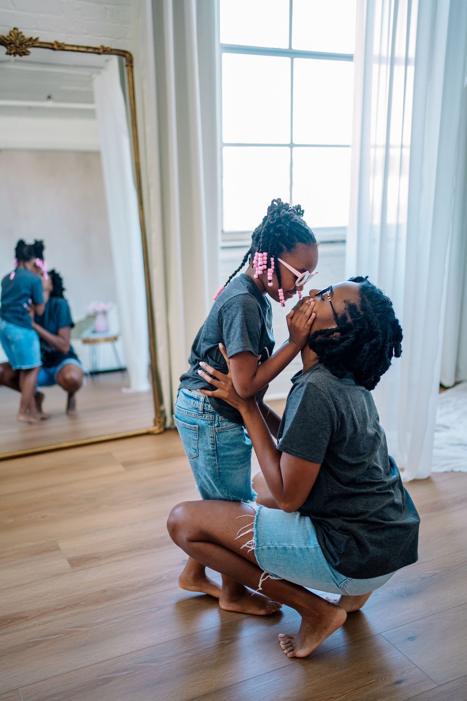 A mother and daughter in matching gray shirts share a tender moment, touching foreheads indoors near a large mirror.