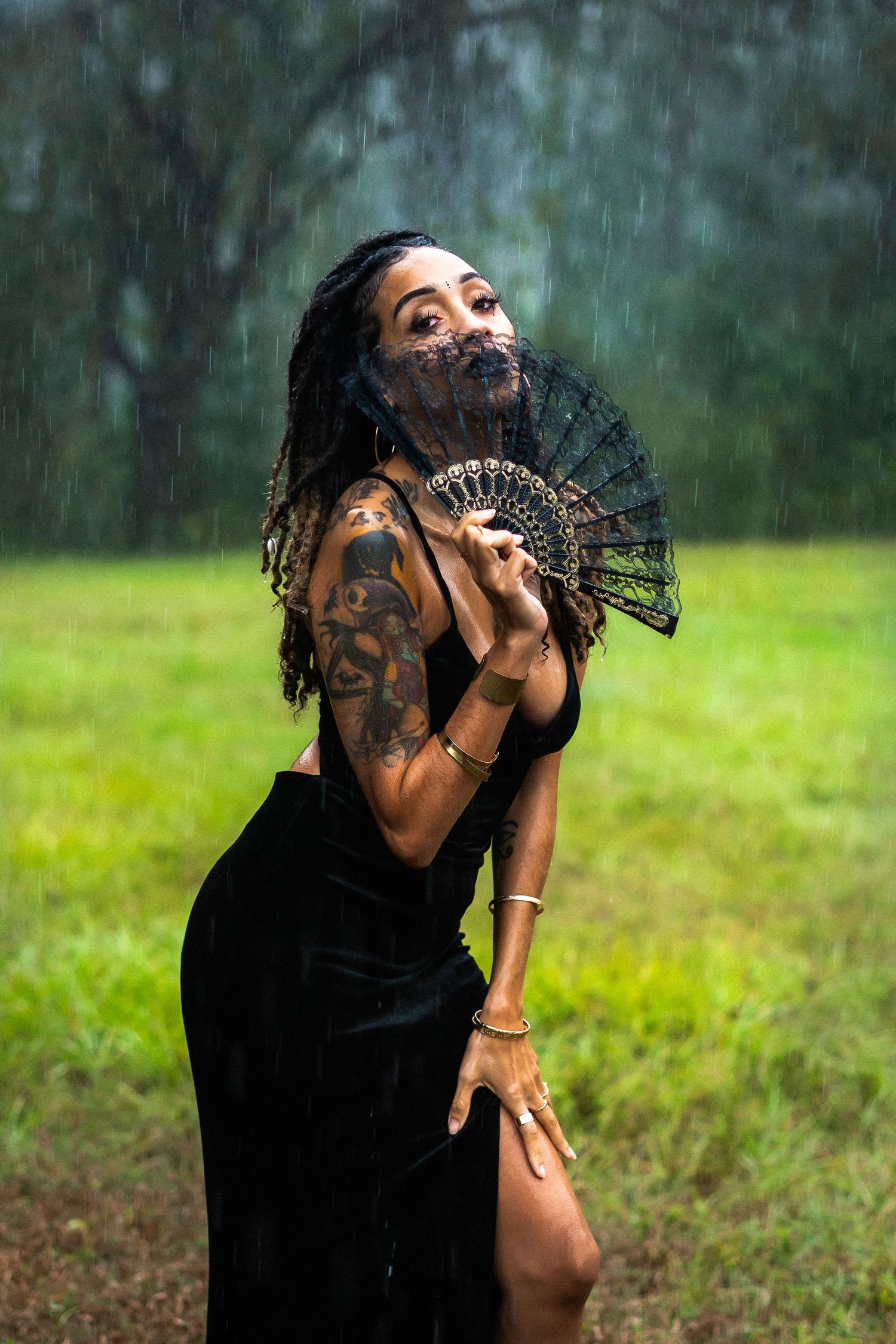 A figure in a black dress dances expressively in a lush green outdoor setting during rainfall.