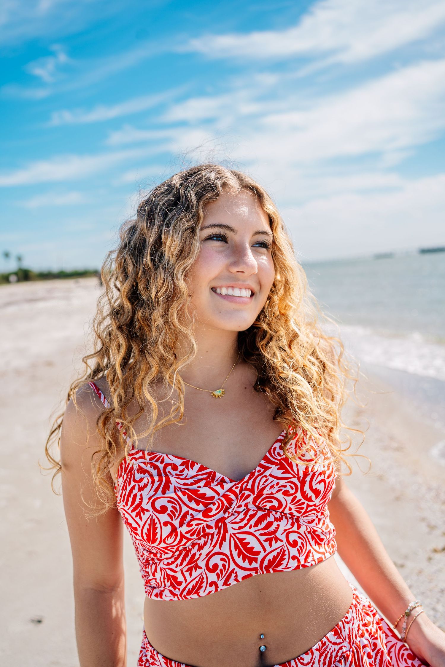 Young woman with curly blonde hair in red floral bikini set smiling on sunny beach by the ocean.