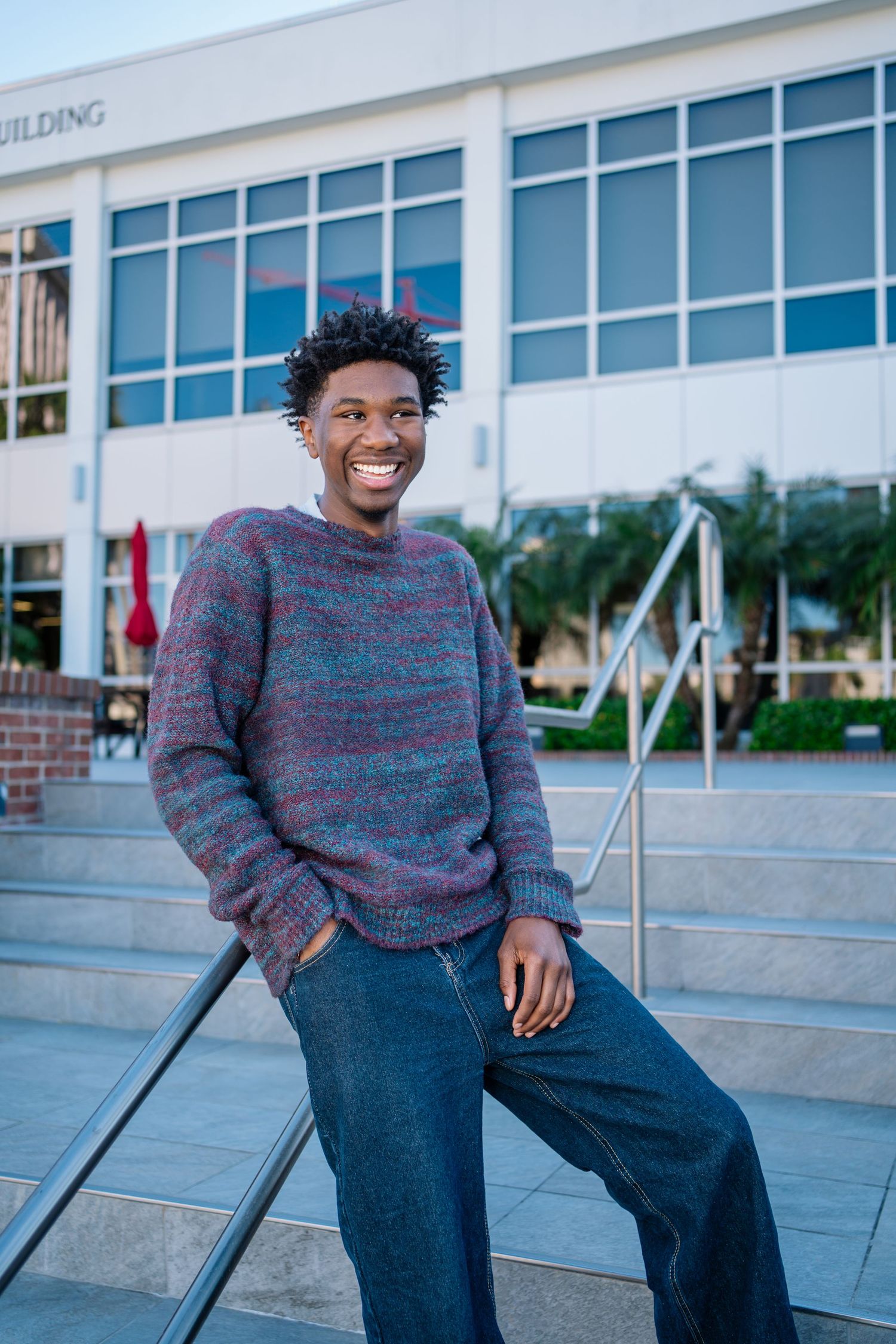 Smiling young Black man in a colorful striped sweater and jeans sitting on steps outside a modern building.
