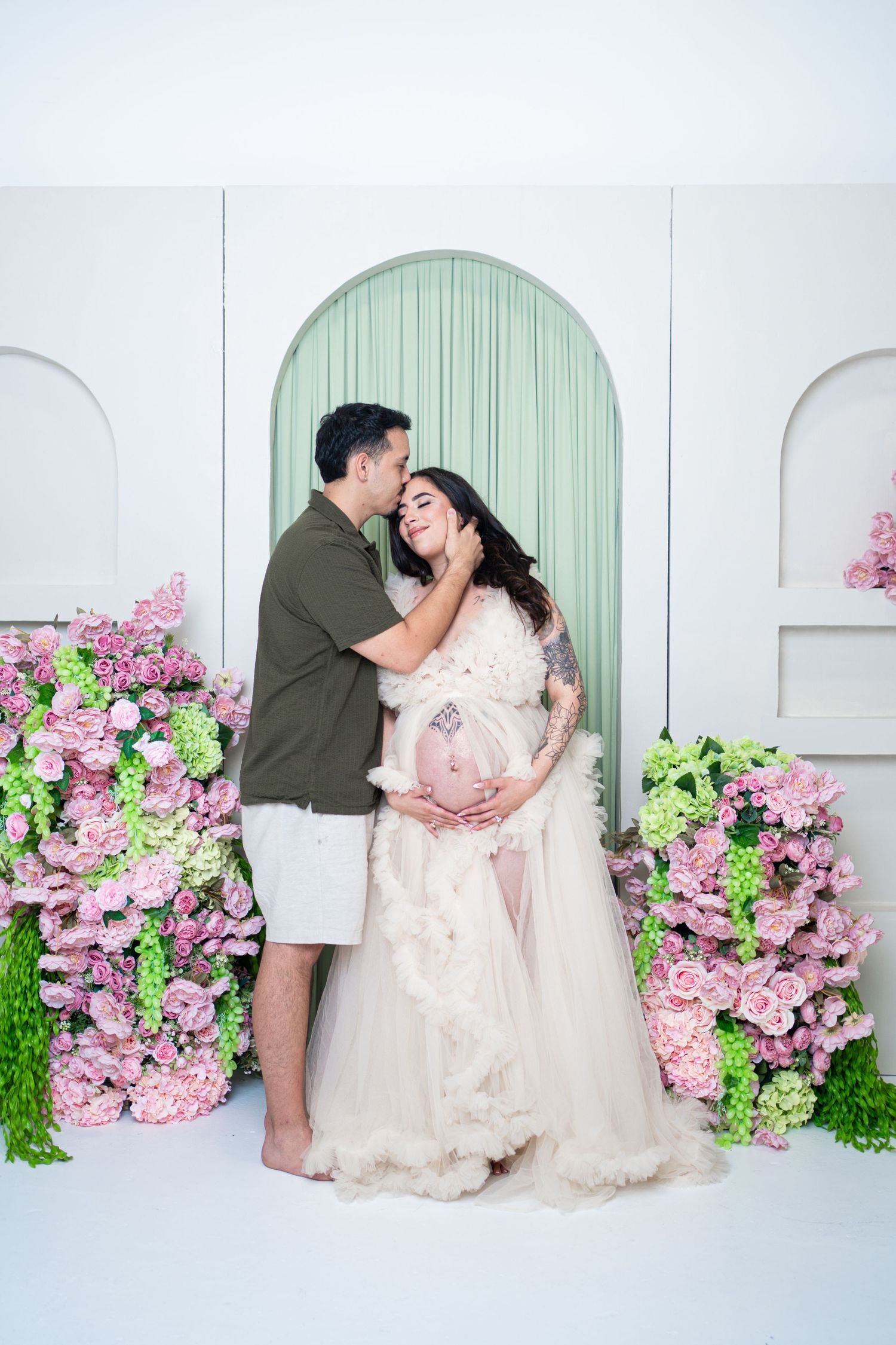 Couple shares tender maternity photo surrounded by pink flowers, woman in white gown cradling baby bump.