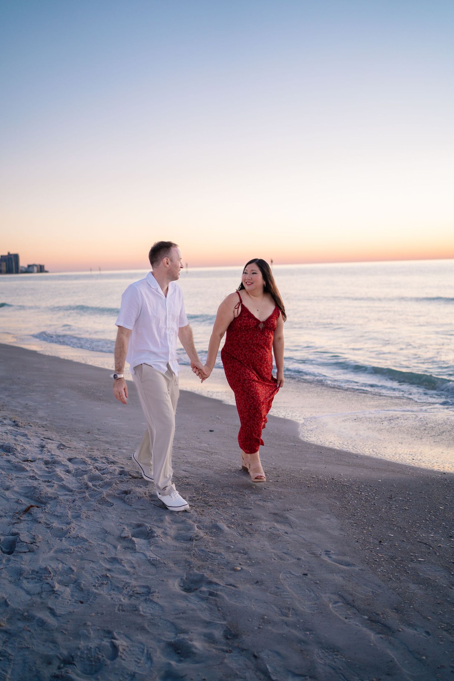 Couple holding hands walking on beach at sunset, woman in red dress and man in white shirt, ocean backdrop.