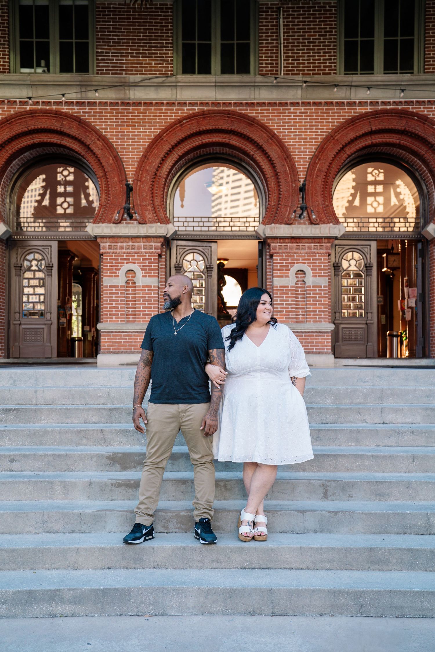 Couple posing on steps of historic red brick building with arched windows, man in blue shirt and woman in white dress.