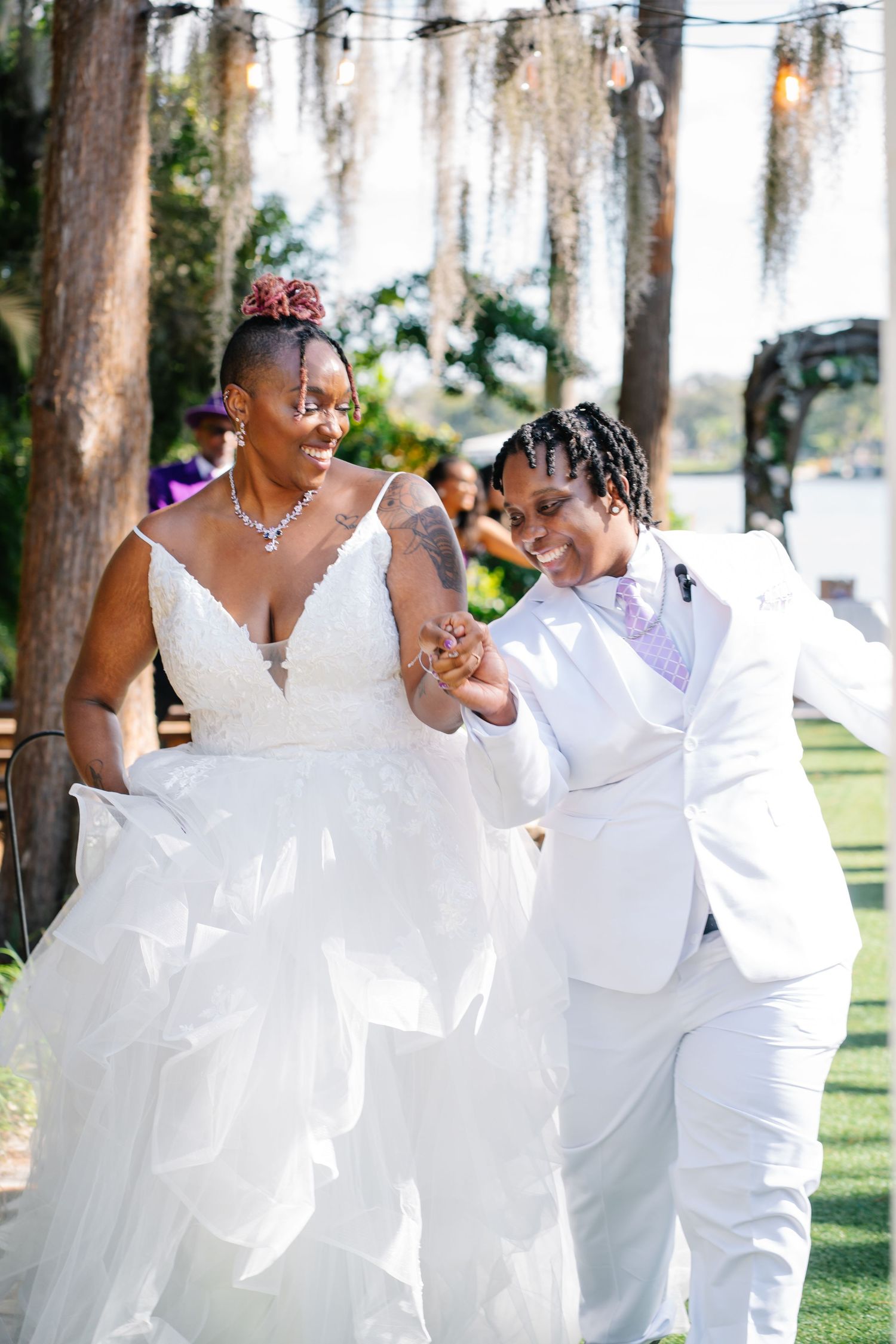 Two brides walk together outdoors, one in a white gown and one in a white suit, smiling at their wedding ceremony.
