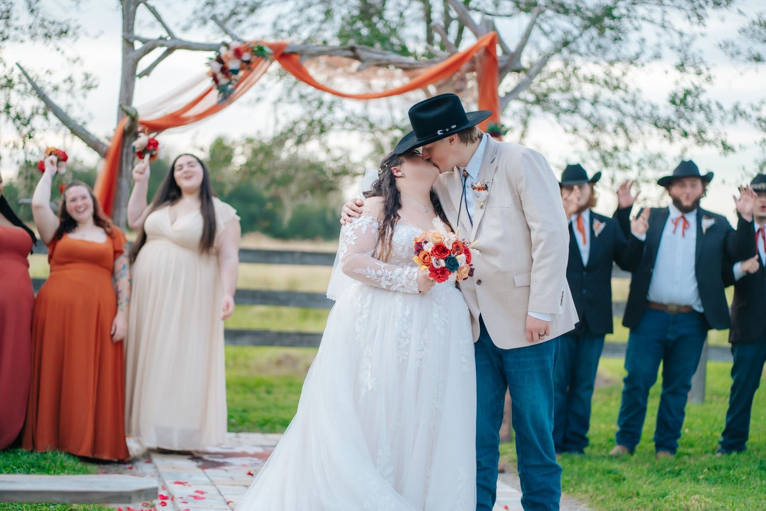 Wedding party in blue suits and orange dresses poses together outdoors under trees.
