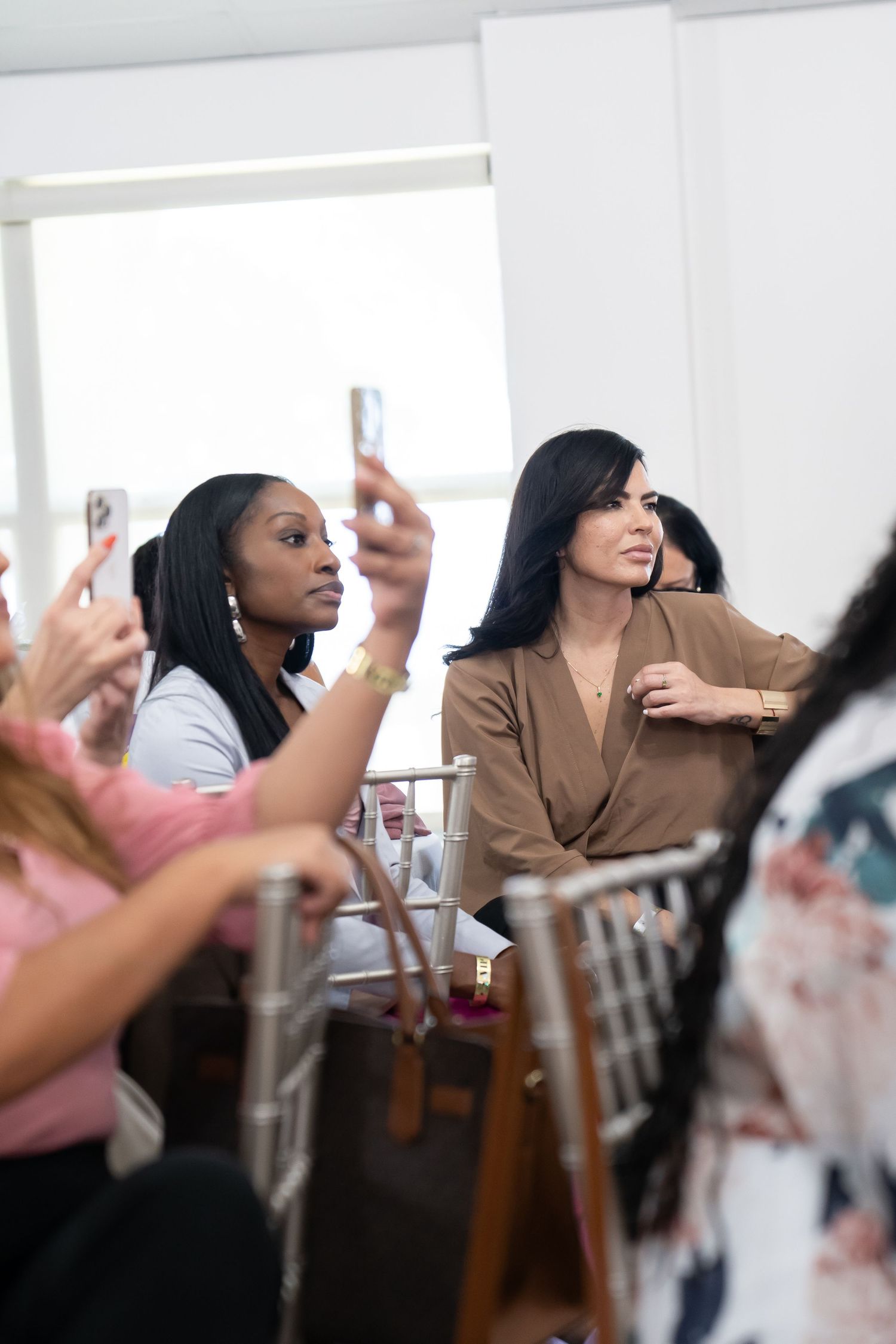 Diverse women seated at event, one taking photo with smartphone, another in brown blazer.