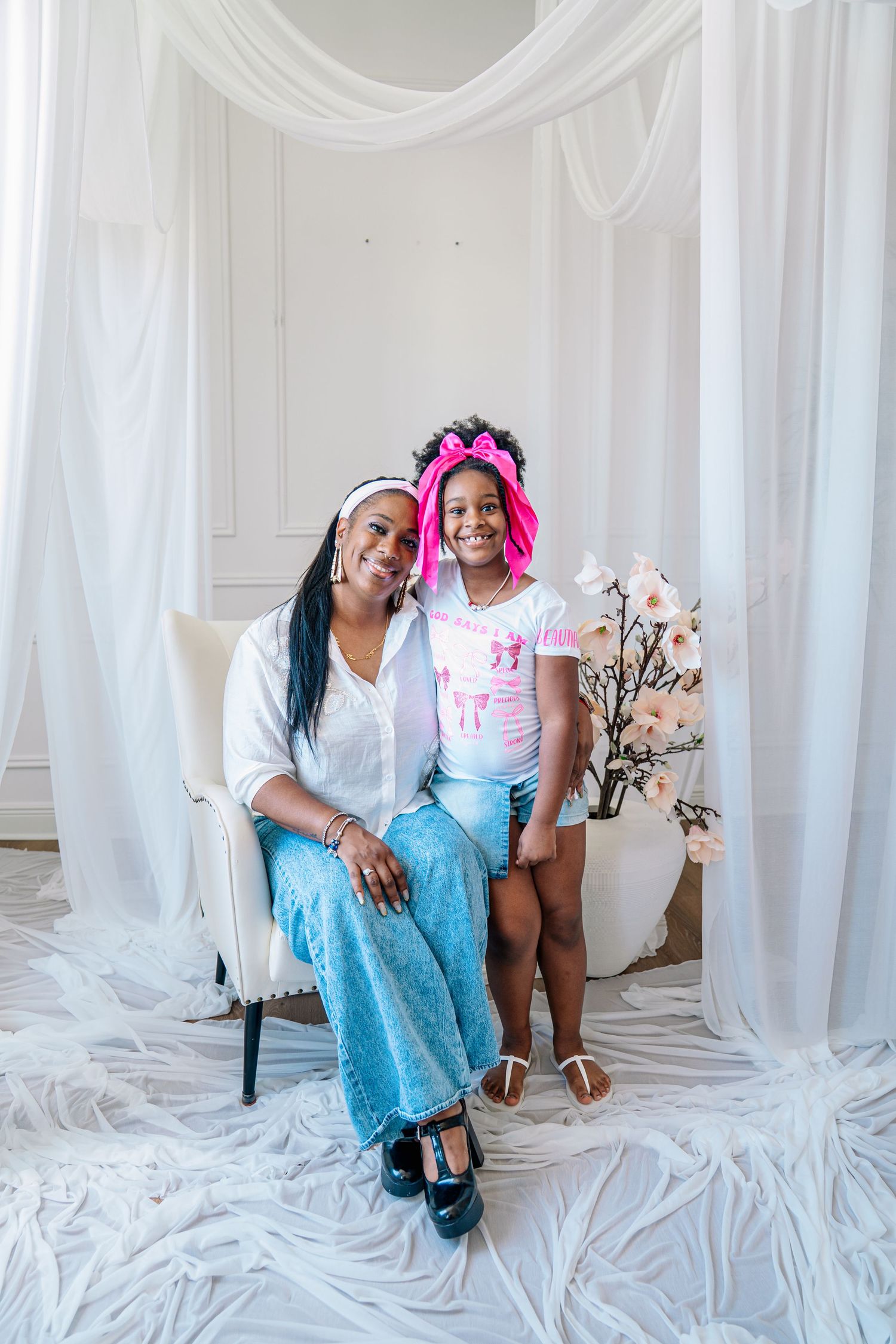 A woman and young girl with a pink bow headband pose together smiling in a bright white draped studio setting.