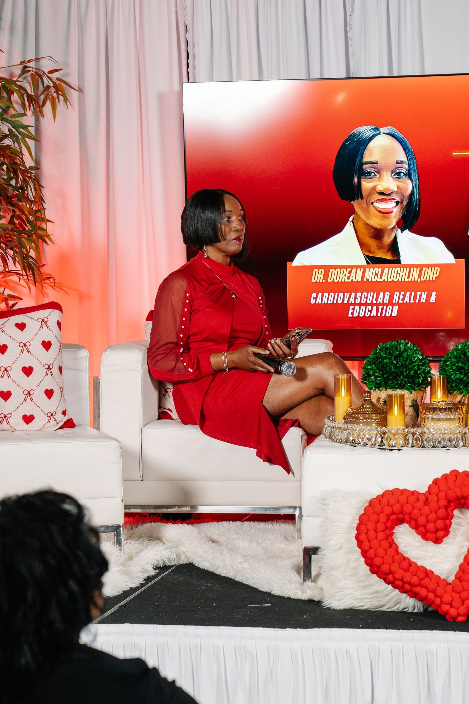 Dr. Doreen McLaughlin DNP speaks on cardiovascular health and education at a red-themed event on a white sofa.
