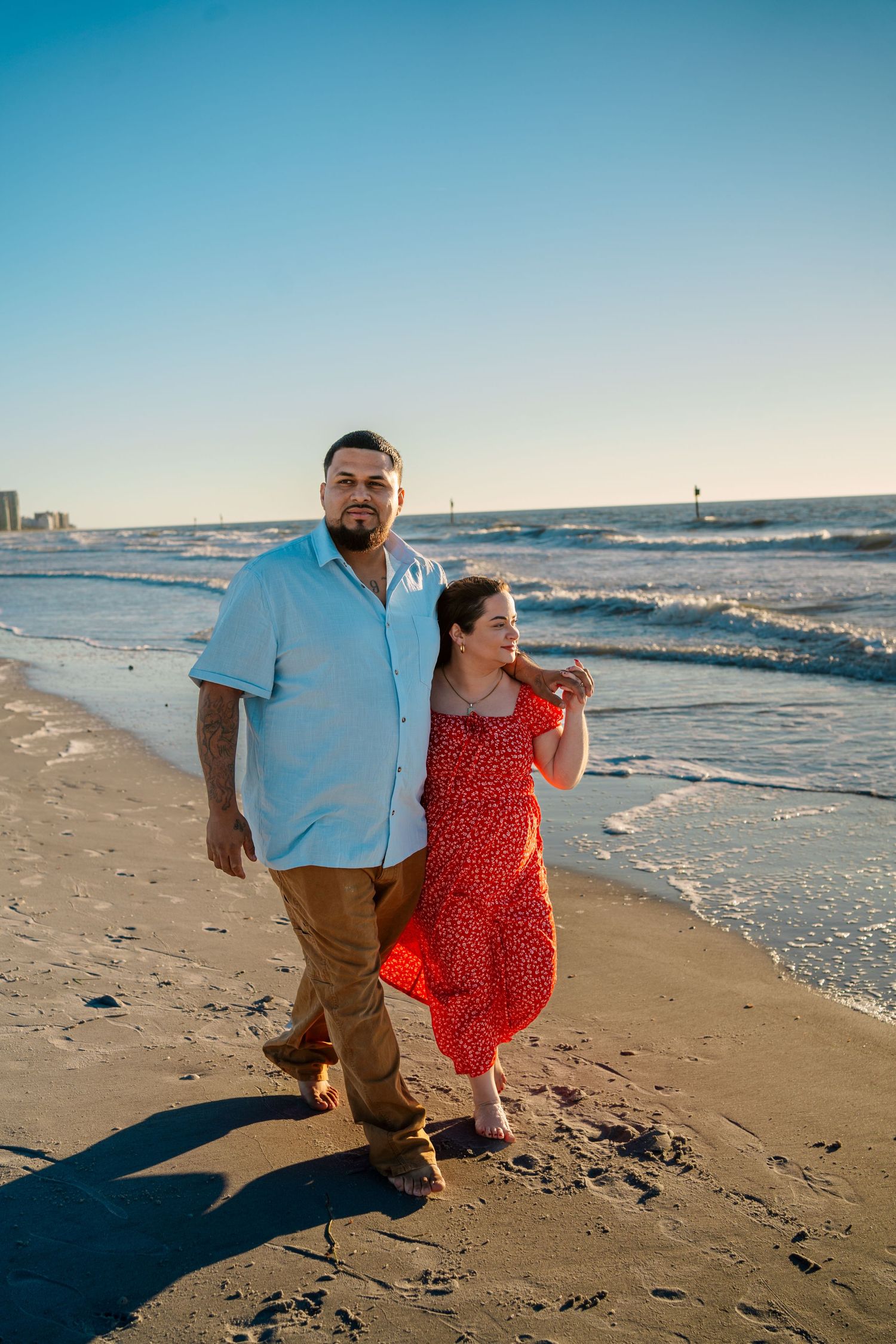 Couple walking hand in hand on a sunny beach, man in blue shirt and woman in red floral dress.