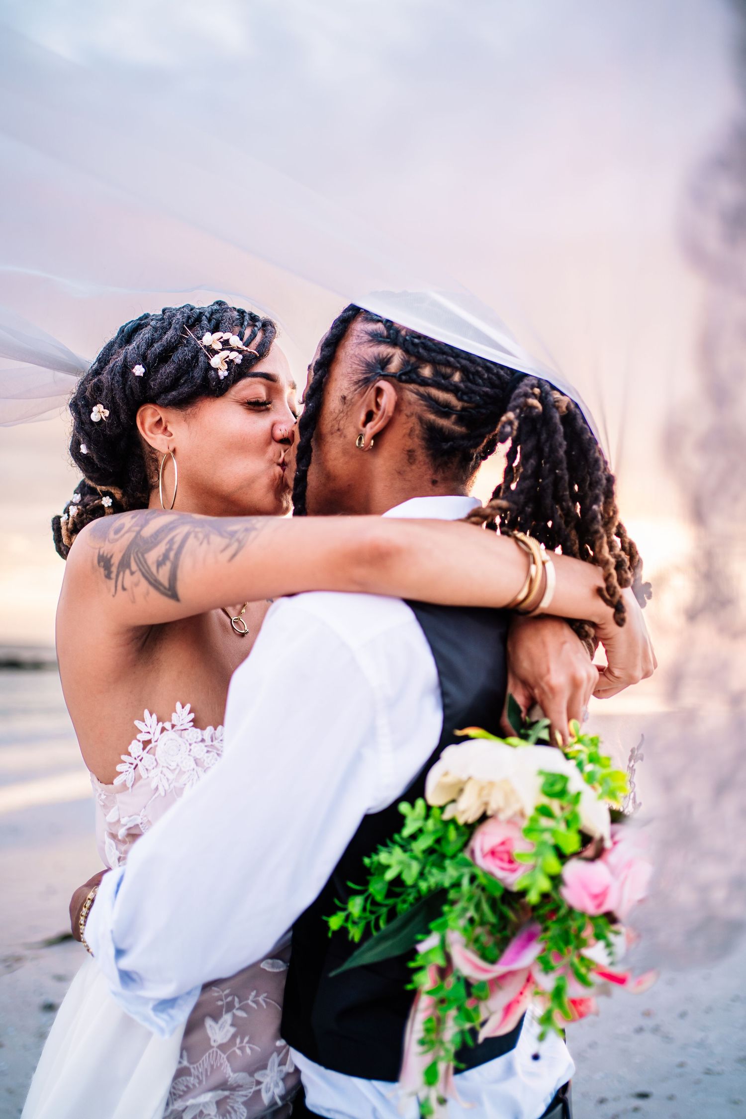 A bride and groom share a romantic kiss under a veil at sunset, with the bride holding a colorful bouquet.