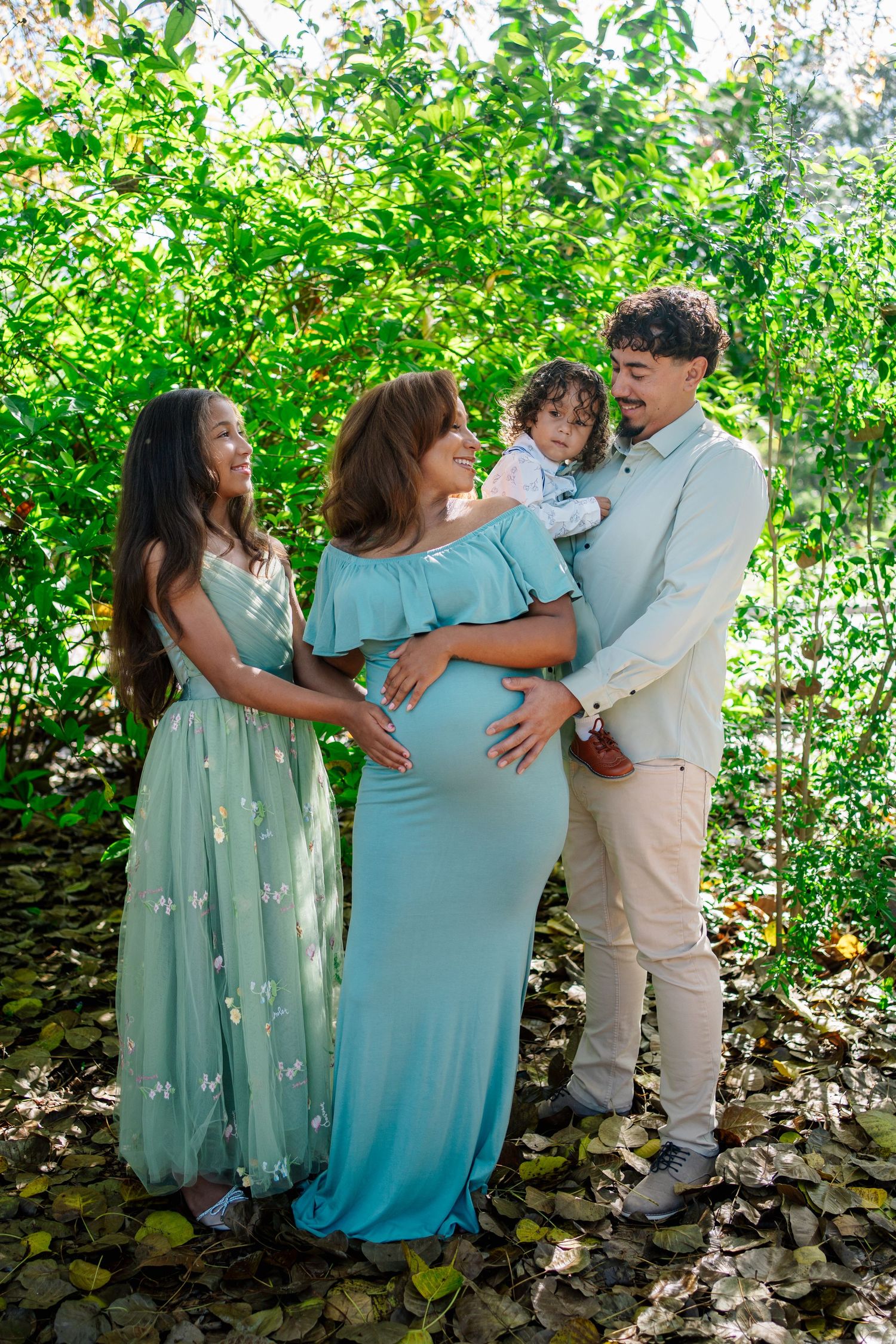 Pregnant woman in teal dress poses with family outdoors among lush green foliage during maternity photo session.