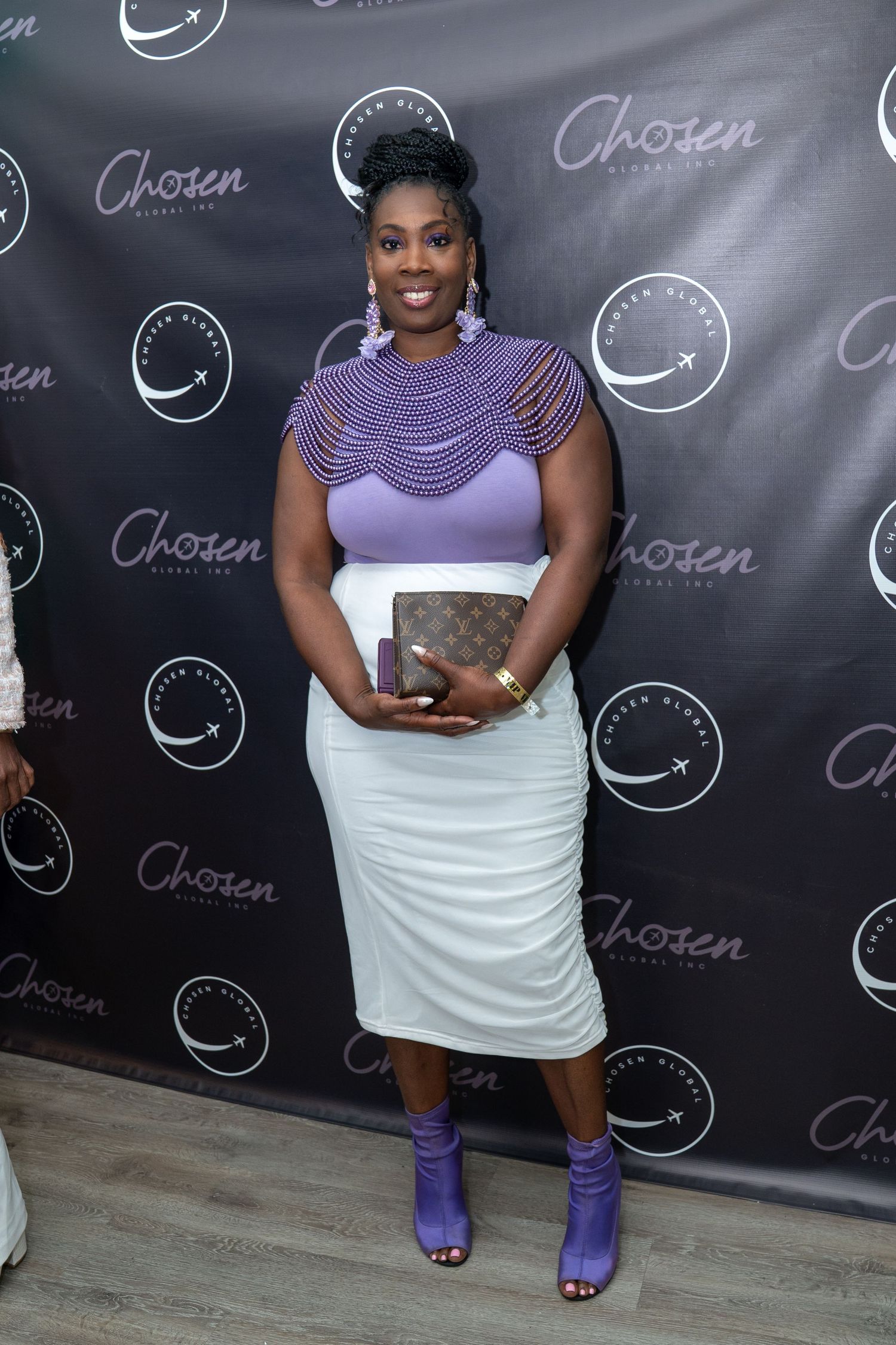 Woman posing in purple beaded top and white skirt at Chosen Global Inc event backdrop.