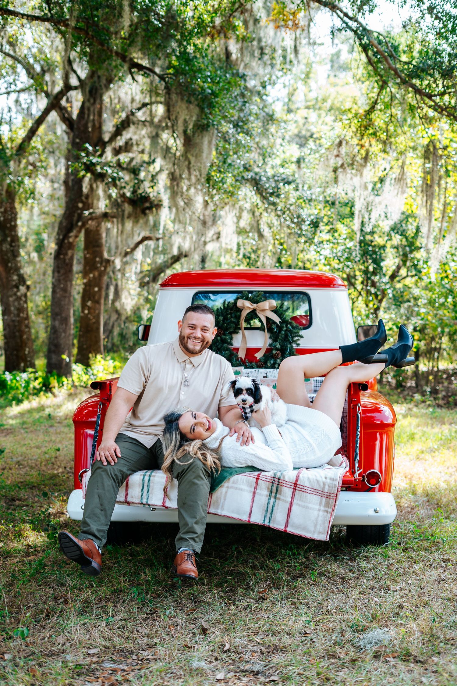 A couple relaxing in the bed of a red pickup truck under trees with hanging moss.