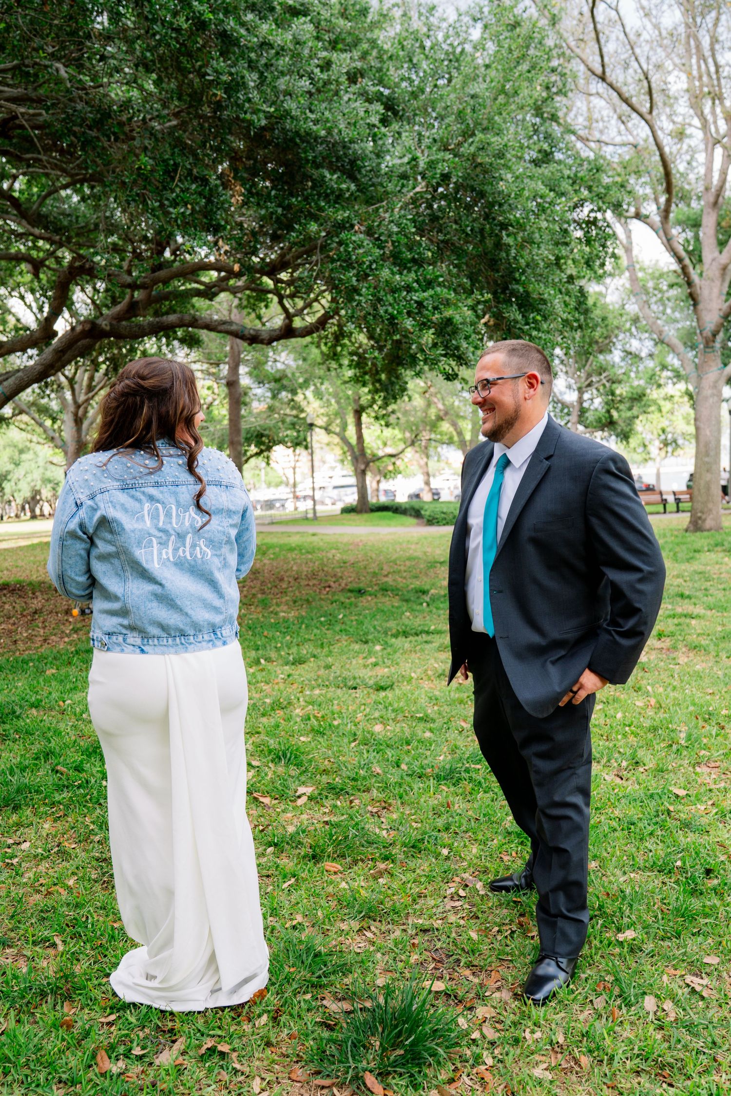 Bride in denim jacket and groom in dark suit share a first look moment in a lush green park.